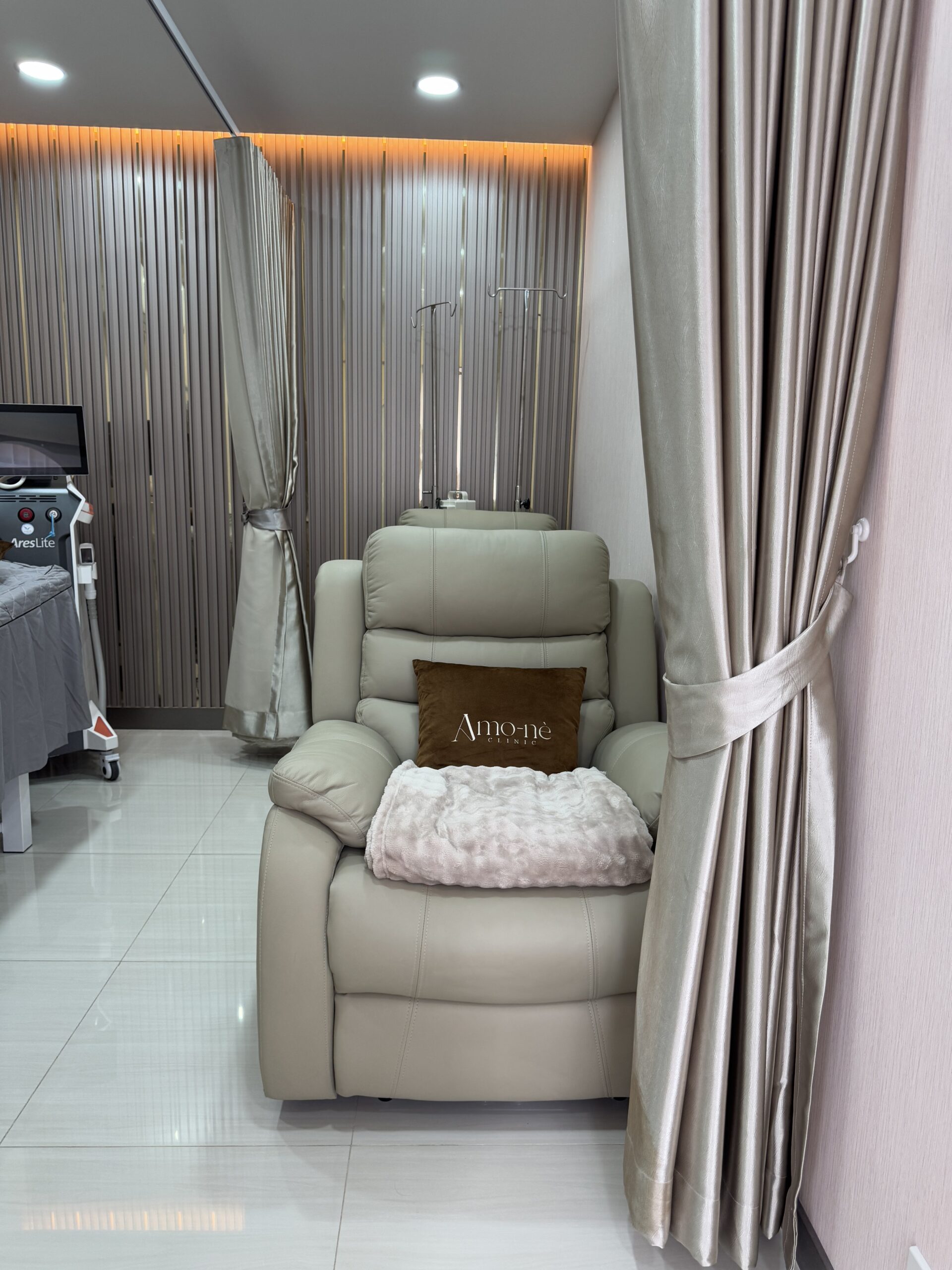 เก้าอี้ปรับเอนหนังสีครีมพร้อมเสาแขวนน้ำเกลือในห้องทรีตเมนต์คลินิก Amo-nè ตกแต่งด้วยผ้าม่านสีเงินและไฟ LED