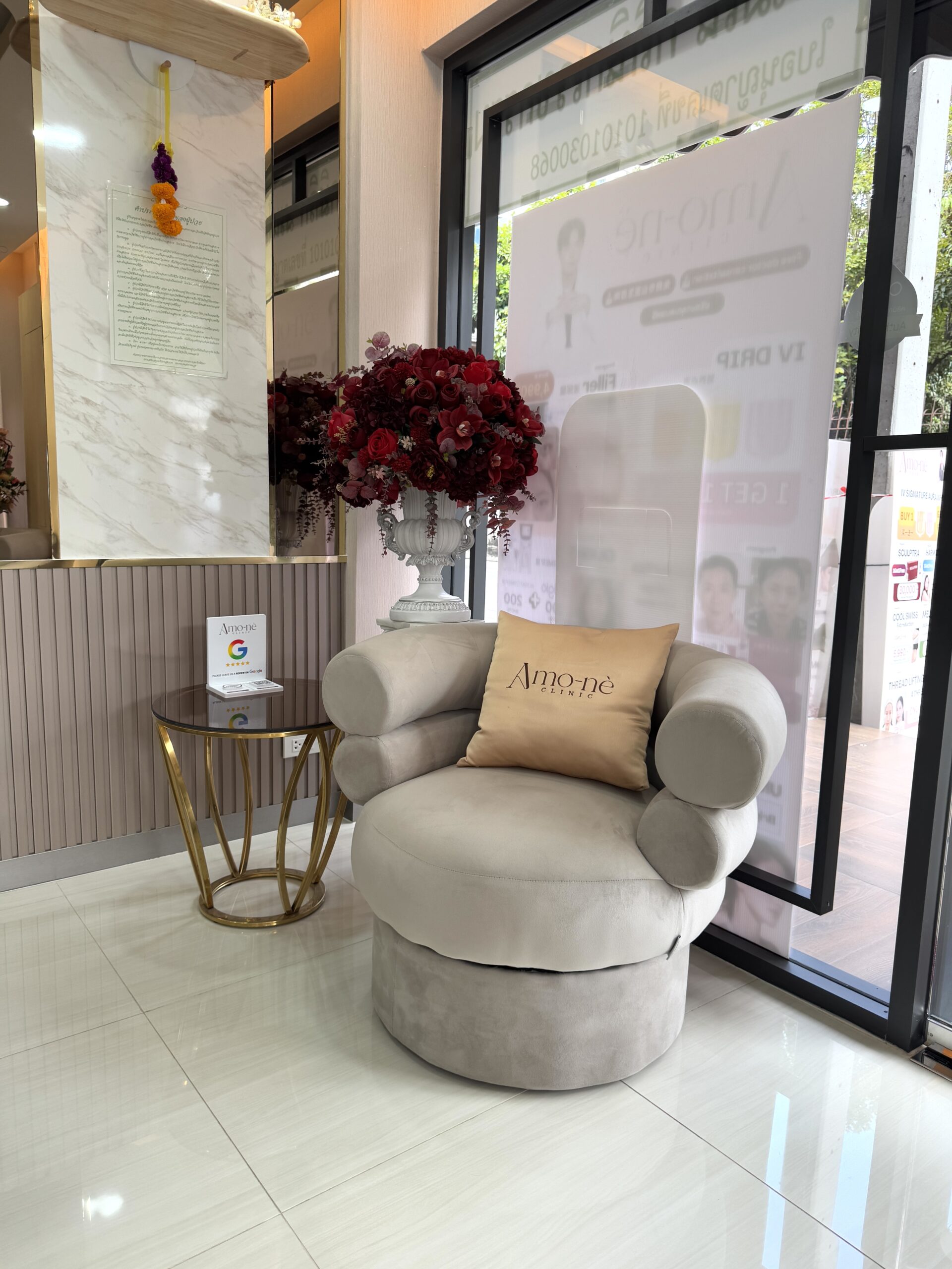 โซฟานั่งสีเทาพร้อมหมอนปักโลโก้ Amo-nè Clinic ในล็อบบี้คลินิกความงามตกแต่งหรูหราพร้อมแจกันดอกไม้สีแดงและโต๊ะข้างสีทอง