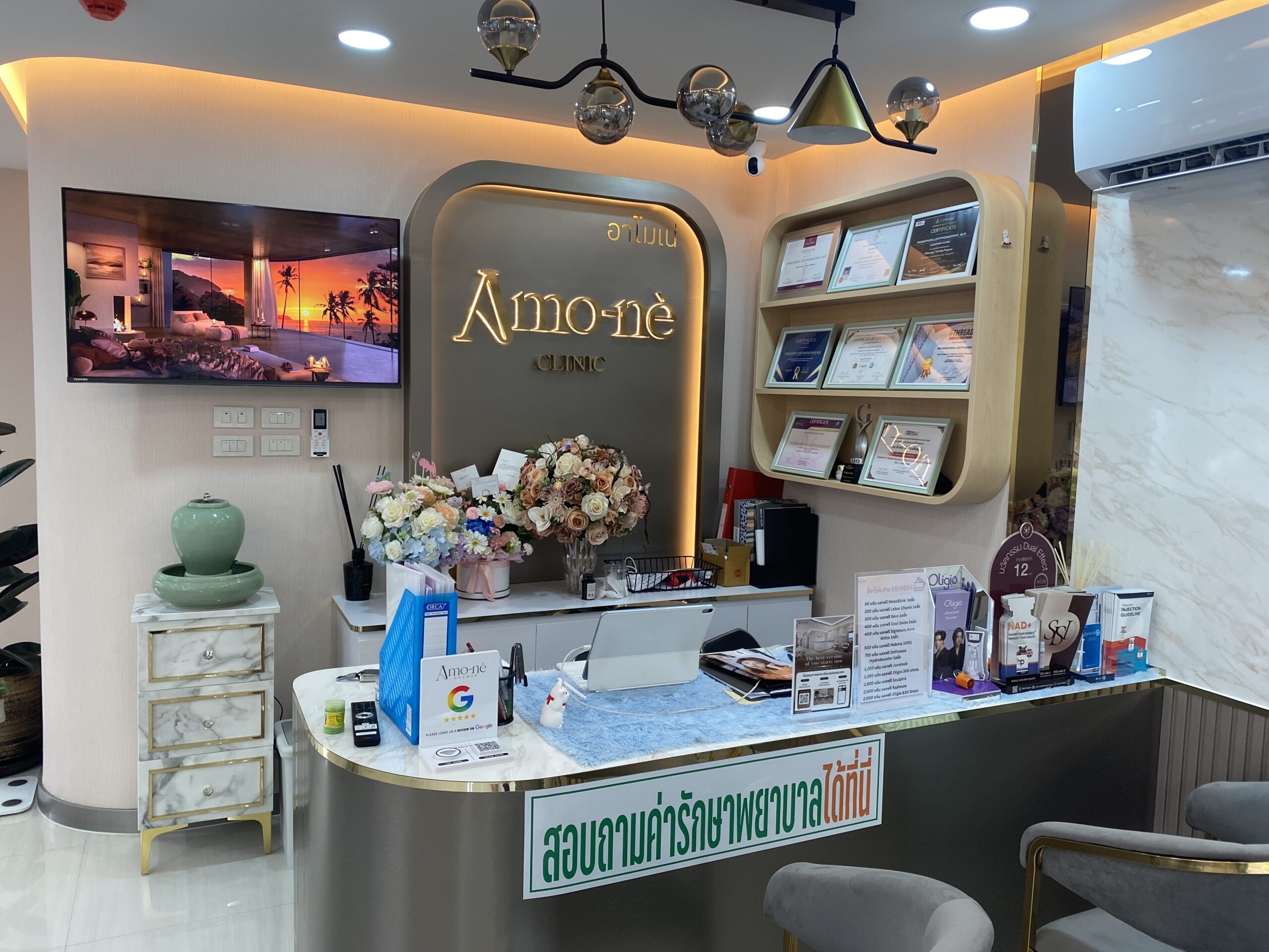 เคาน์เตอร์ต้อนรับของคลินิกอาโมเน่ ตกแต่งหรูหราพร้อมป้ายชื่อไฟ LED ดอกไม้ประดับ และใบประกาศนียบัตรบนชั้นวาง