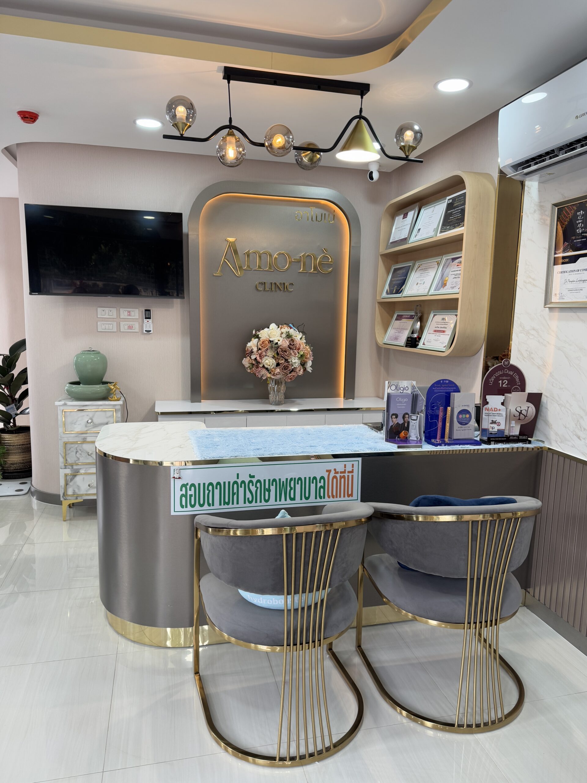 เคาน์เตอร์ต้อนรับของคลินิก Amo-né ตกแต่งหรูหราด้วยโทนสีเทาและทอง มีเก้าอี้กำมะหยี่สีเทาและป้ายสอบถามค่ารักษาพยาบาล