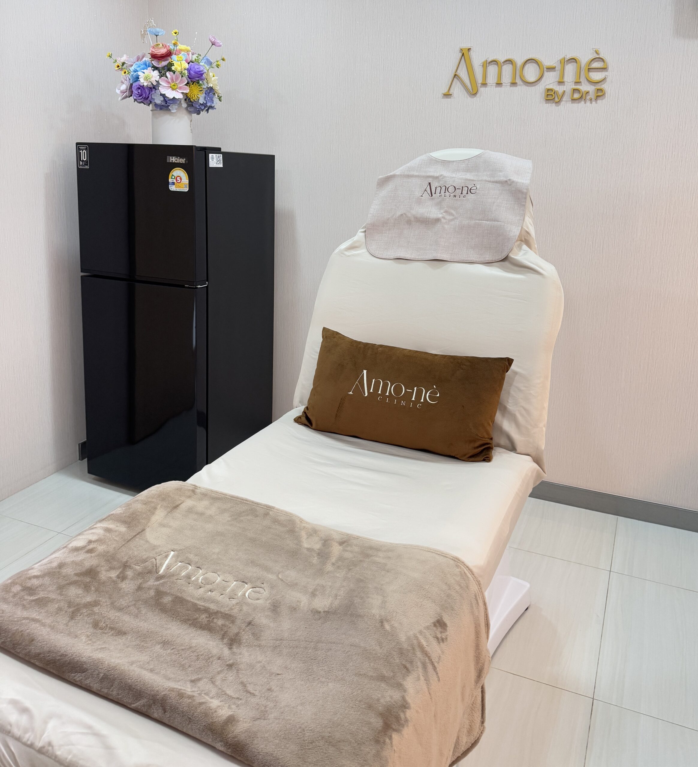 ห้องทรีตเมนต์คลินิก Amo-nè By Dr.P พร้อมเตียงนวดสีขาว หมอนและผ้าห่มสีน้ำตาล ตู้เย็น Haier สีดำ และแจกันดอกไม้