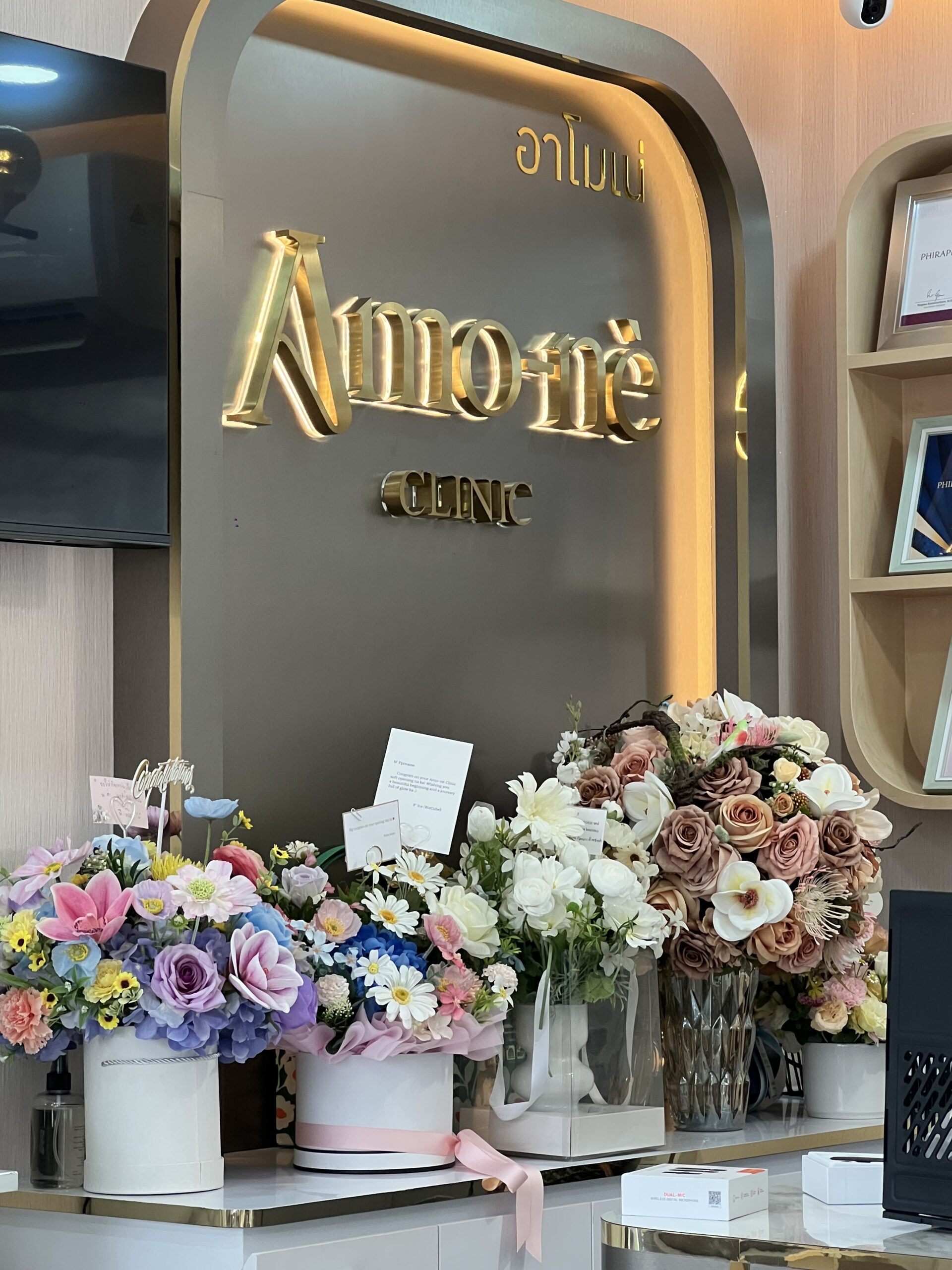 บริเวณแผนกต้อนรับของคลินิกอาโมเน่ ตกแต่งด้วยช่อดอกไม้หลากสีสันสวยงาม พร้อมป้ายชื่อตัวอักษรสีทองบนพื้นหลังสีเทา