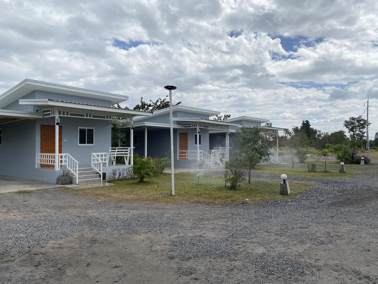 บังกะโลสีฟ้าหลายหลังในรีสอร์ทชนบท มีบันไดสีขาว ประตูไม้สีน้ำตาล สนามหญ้าและทางกรวด ท้องฟ้ามีเมฆ