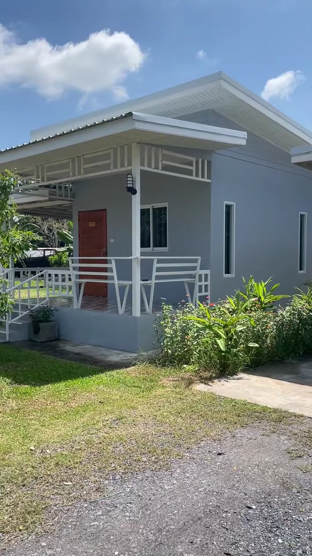 บ้านพักขนาดเล็กสีเทาอ่อนหลังคาจั่วสีขาวมีระเบียงหน้าบ้านพร้อมสวนสีเขียว