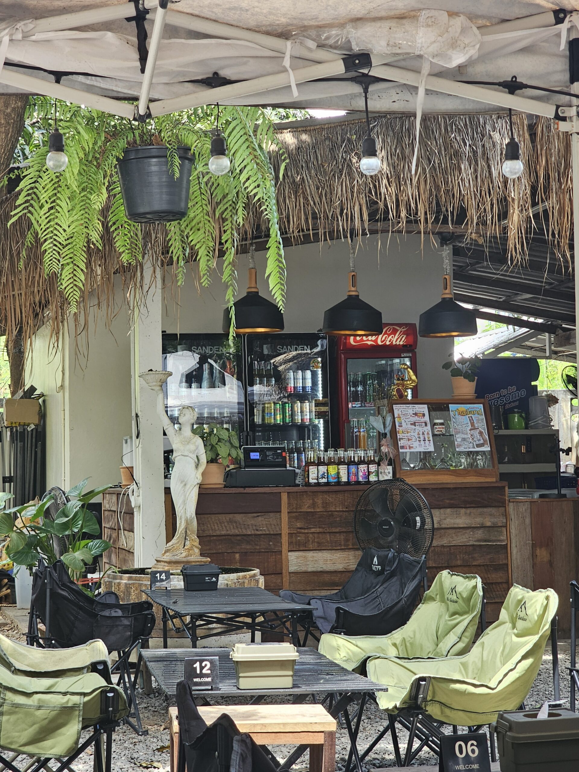 บาร์กลางแจ้งสไตล์แคมป์ปิ้งพร้อมเก้าอี้สนามสีเขียว ตู้เย็นโคคา-โคล่า โคมไฟแขวน เฟิร์นห้อย และรูปปั้นสีขาวหน้าเคาน์เตอร์ไม้