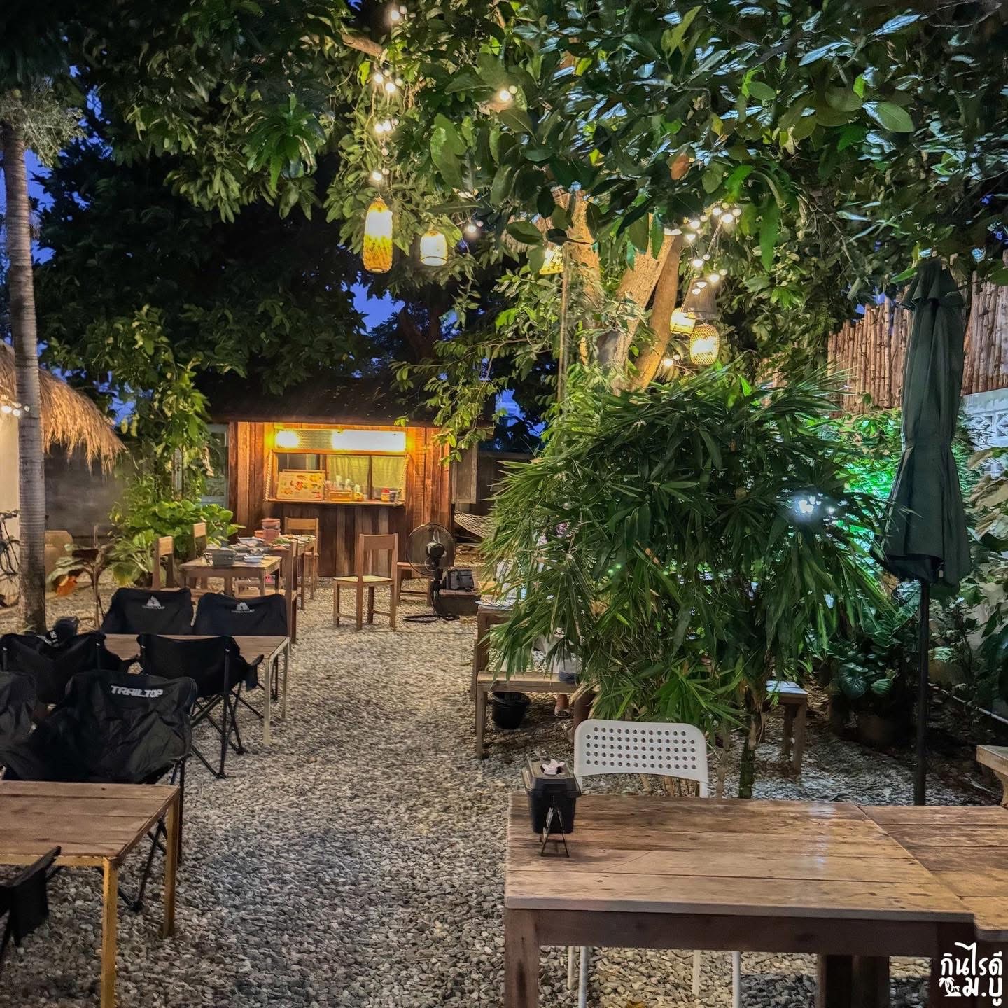 บรรยากาศร้านอาหารสวนกลางแจ้งยามเย็น มีโต๊ะไม้ เก้าอี้แคมป์ ไฟประดับบนต้นไม้ใหญ่ และร้านค้าเล็กๆ สไตล์กระท่อมไม้