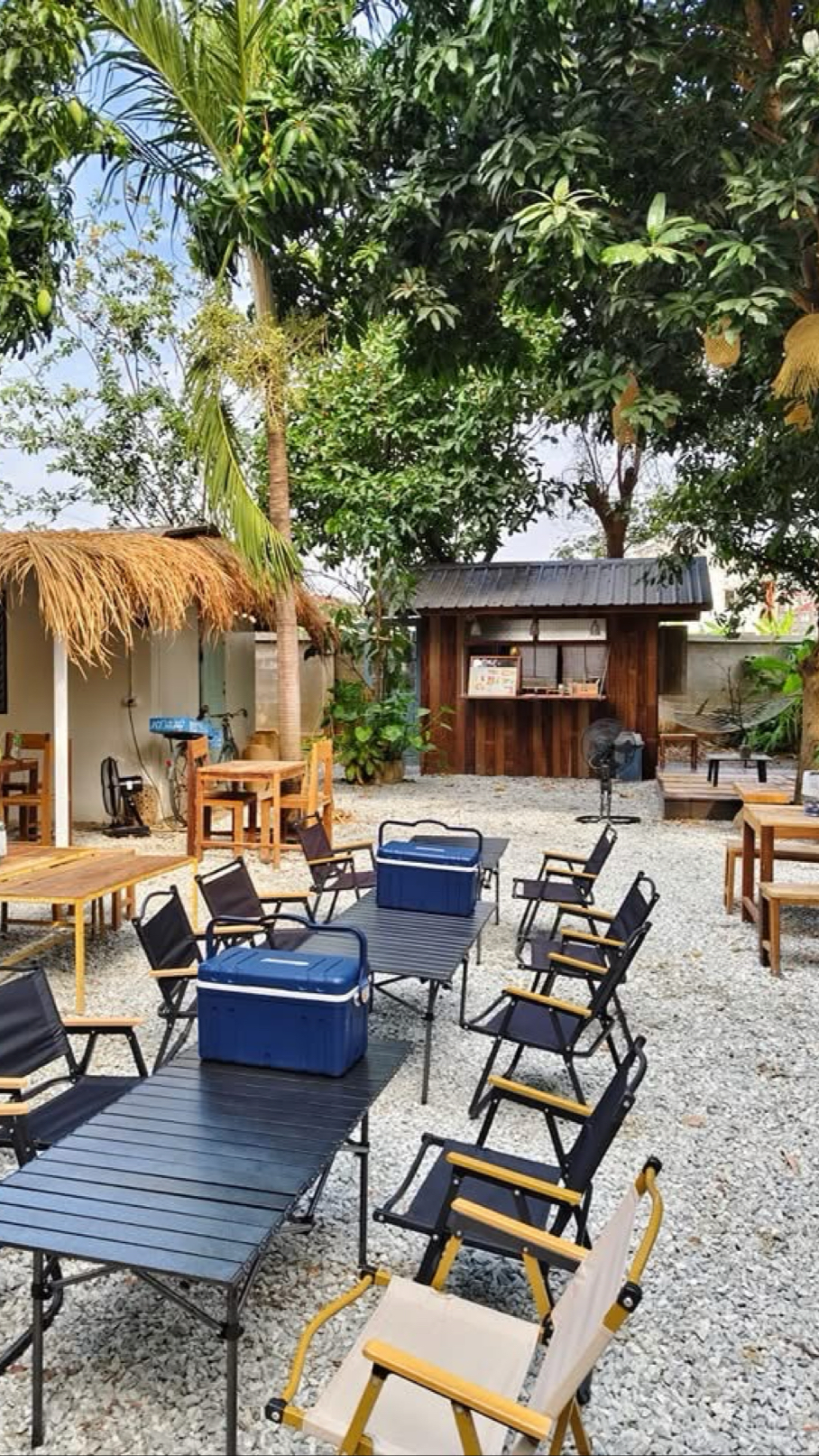 พื้นที่นั่งกลางแจ้งสไตล์แคมป์ปิ้งพร้อมเก้าอี้พับ โต๊ะพับ กล่องเย็นสีน้ำเงิน และกระท่อมไม้ท่ามกลางสวนร่มรื่น