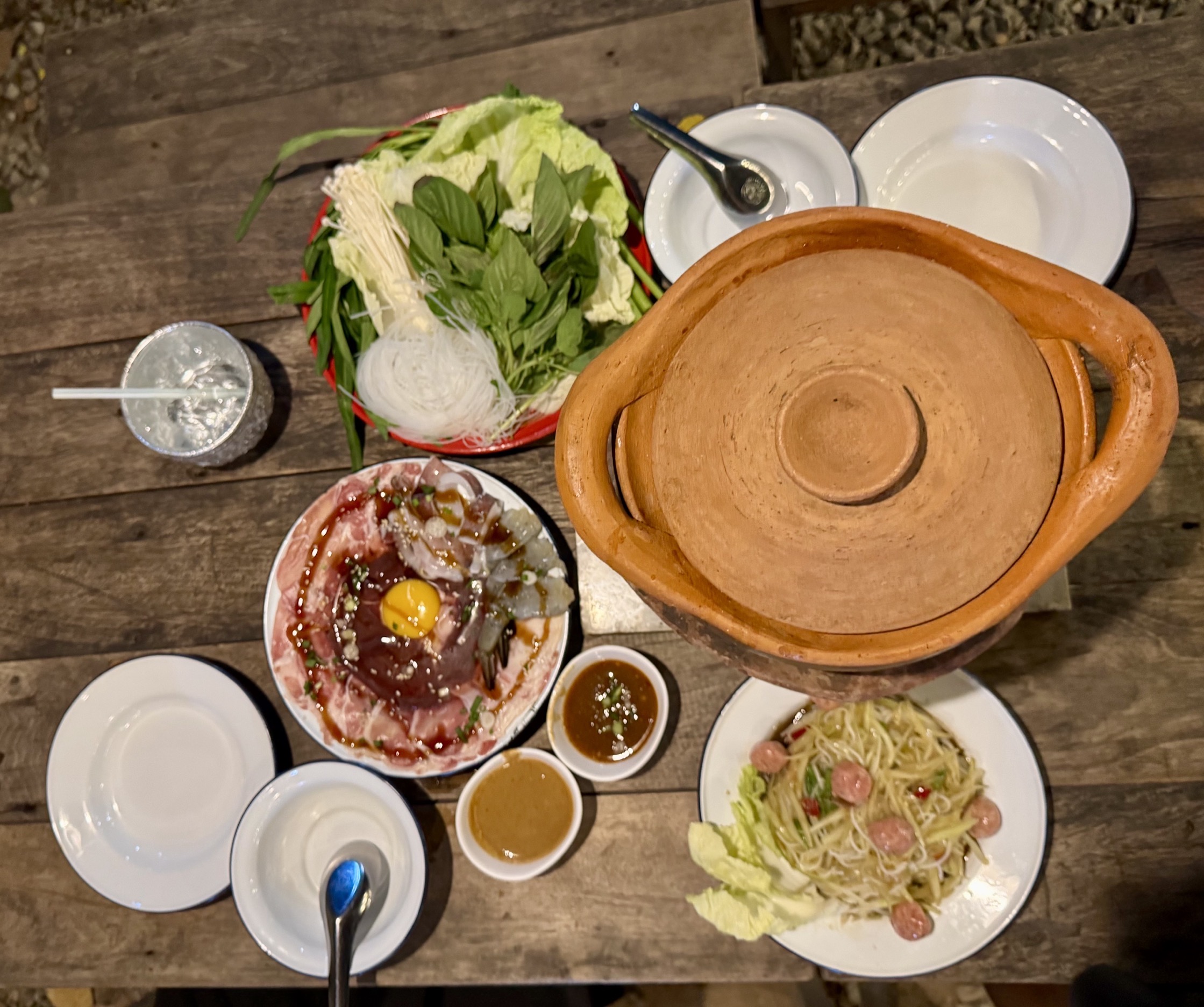 ชุดจิ้มจุ่มหม้อดินพร้อมจานเนื้อสด ผักสด เส้นหมี่ ส้มตำ และน้ำจิ้มบนโต๊ะไม้