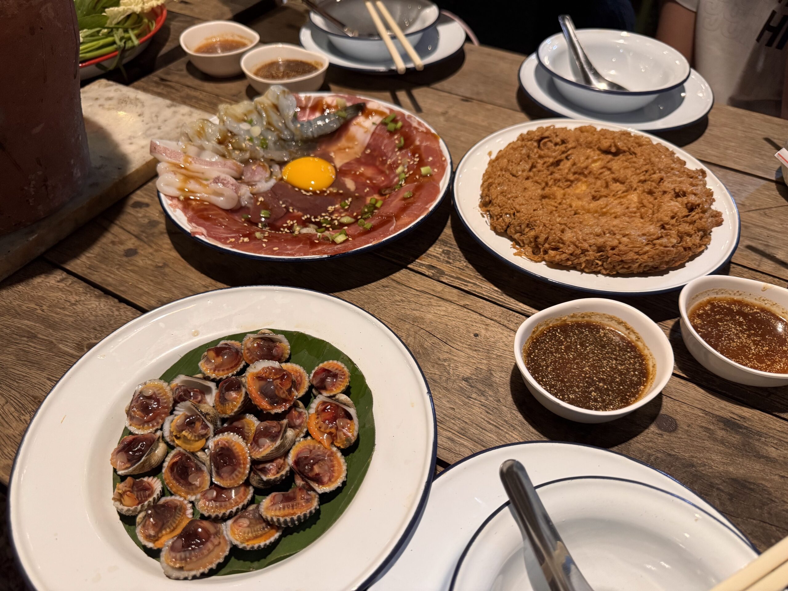 โต๊ะอาหารทะเลย่างสไตล์ไทย มีหอยแครงลวก จานเนื้อและกุ้งดิบพร้อมไข่แดง บะหมี่กรอบ และน้ำจิ้มซีฟู้ดหลายถ้วยบนโต๊ะไม้