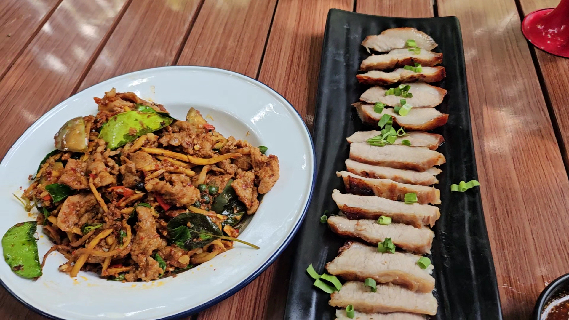 จานผัดเผ็ดหมูกับสมุนไพรบนจานขาวและหมูกรอบหั่นชิ้นโรยต้นหอมบนจานดำวางบนโต๊ะไม้