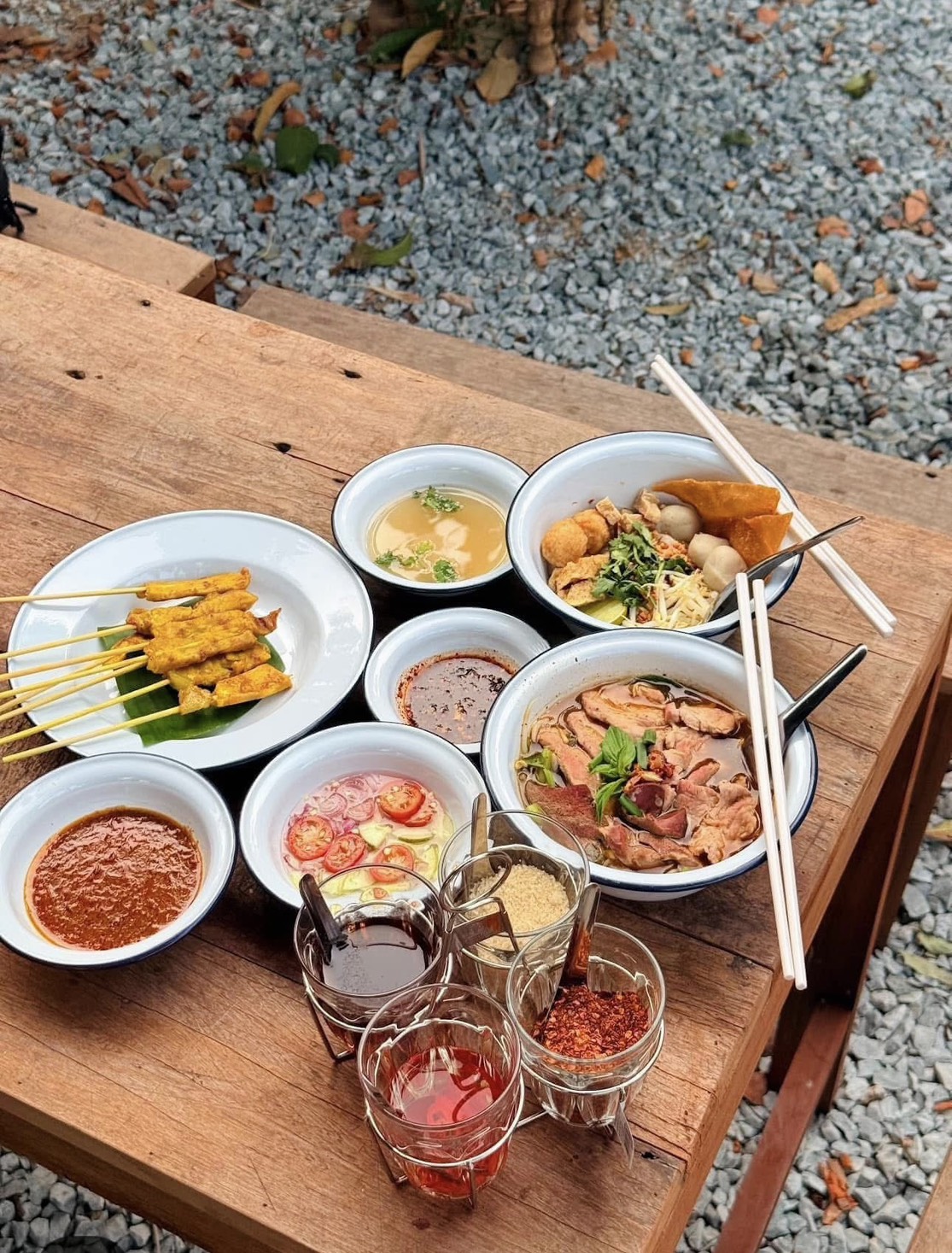 โต๊ะไม้เสิร์ฟอาหารไทยหลากหลาย ได้แก่ หมูสะเต๊ะ ก๋วยเตี๋ยวเนื้อ ก๋วยเตี๋ยวลูกชิ้น น้ำซุป สลัดแตงกวามะเขือเทศ น้ำจิ้มสะเต๊ะ และเครื่องปรุงรสในแก้ว