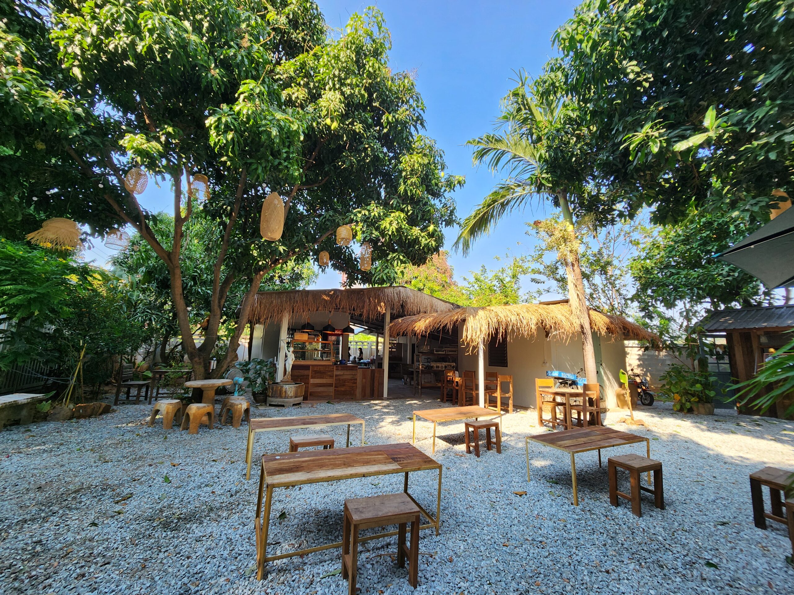 คาเฟ่กลางแจ้งสไตล์ทรอปิคอลพร้อมโต๊ะเก้าอี้ไม้บนพื้นกรวด หลังคามุงจาก ร่มเงาต้นไม้ใหญ่ และโคมไฟแขวนตกแต่ง