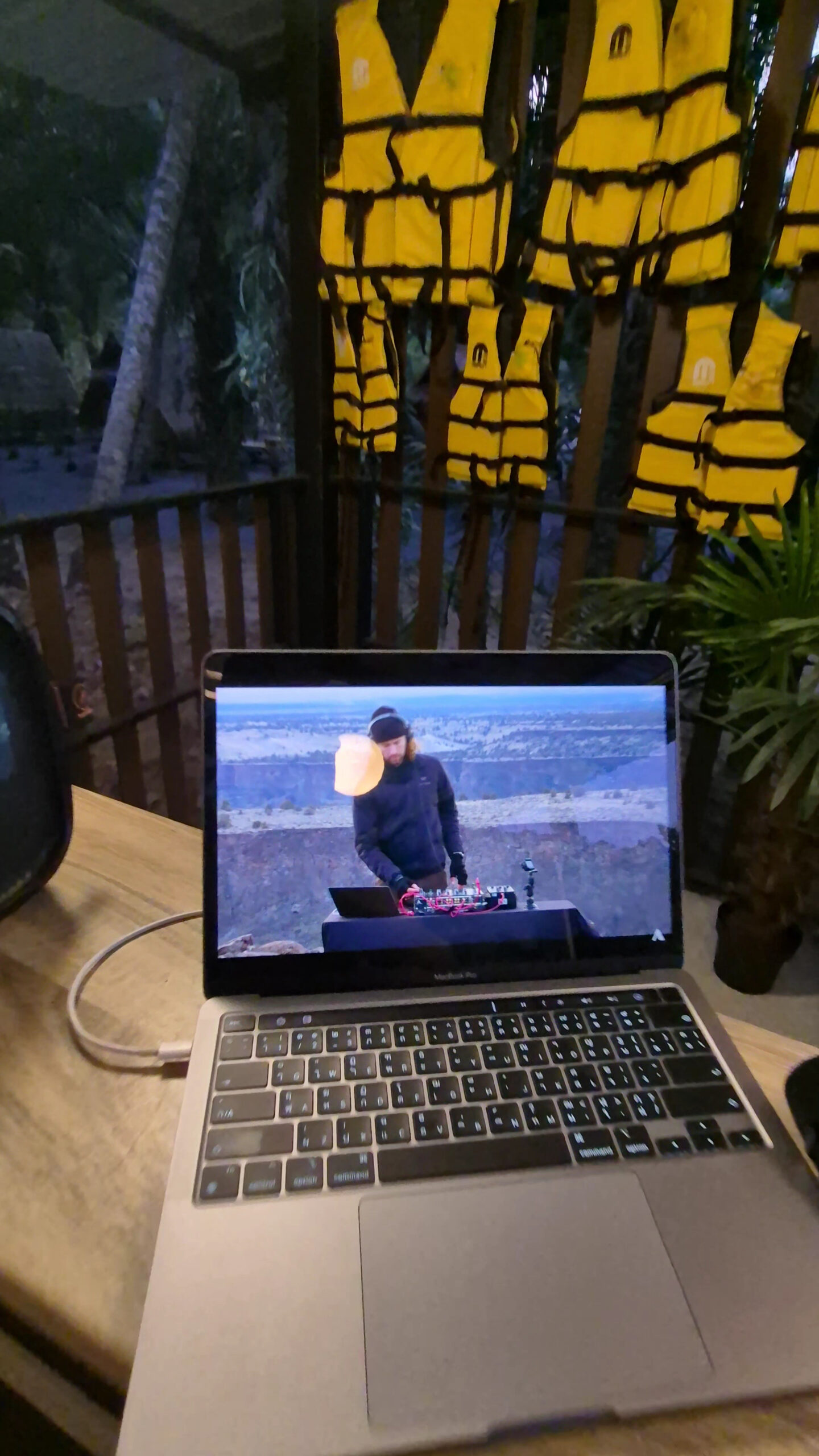 แล็ปท็อป MacBook Pro เปิดวิดีโอดีเจเล่นเพลงบนหน้าผา วางบนโต๊ะไม้ มีเสื้อชูชีพสีเหลืองแขวนอยู่ด้านหลัง บรรยากาศริมน้ำยามค่ำคืน