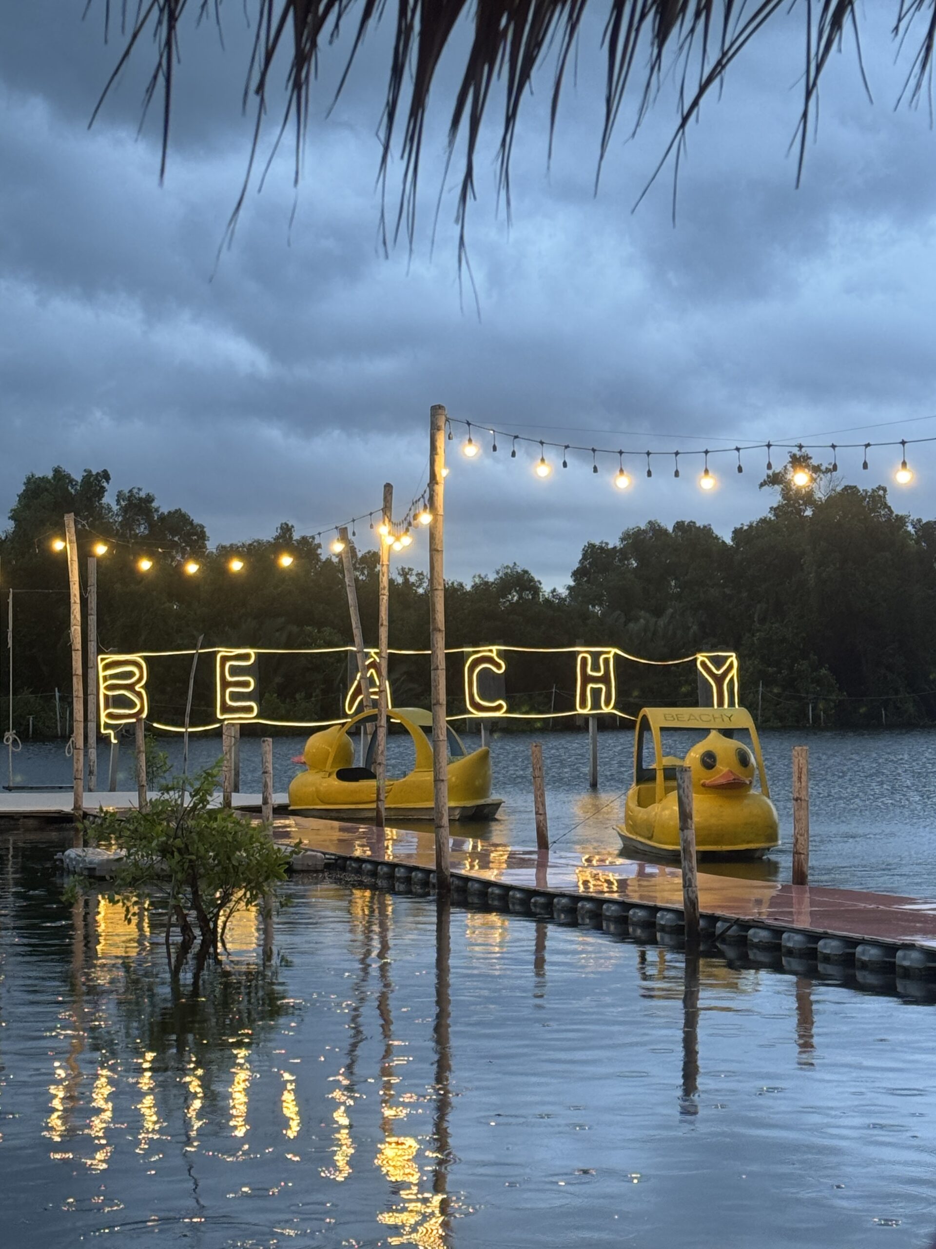 ท่าเรือริมทะเลสาบร้าน Beachy ยามเย็น มีป้ายไฟนีออนสีเหลืองเขียนว่า BEACHY เรือเป็ดสีเหลืองจอดอยู่ และไฟสายประดับบรรยากาศสวยงาม