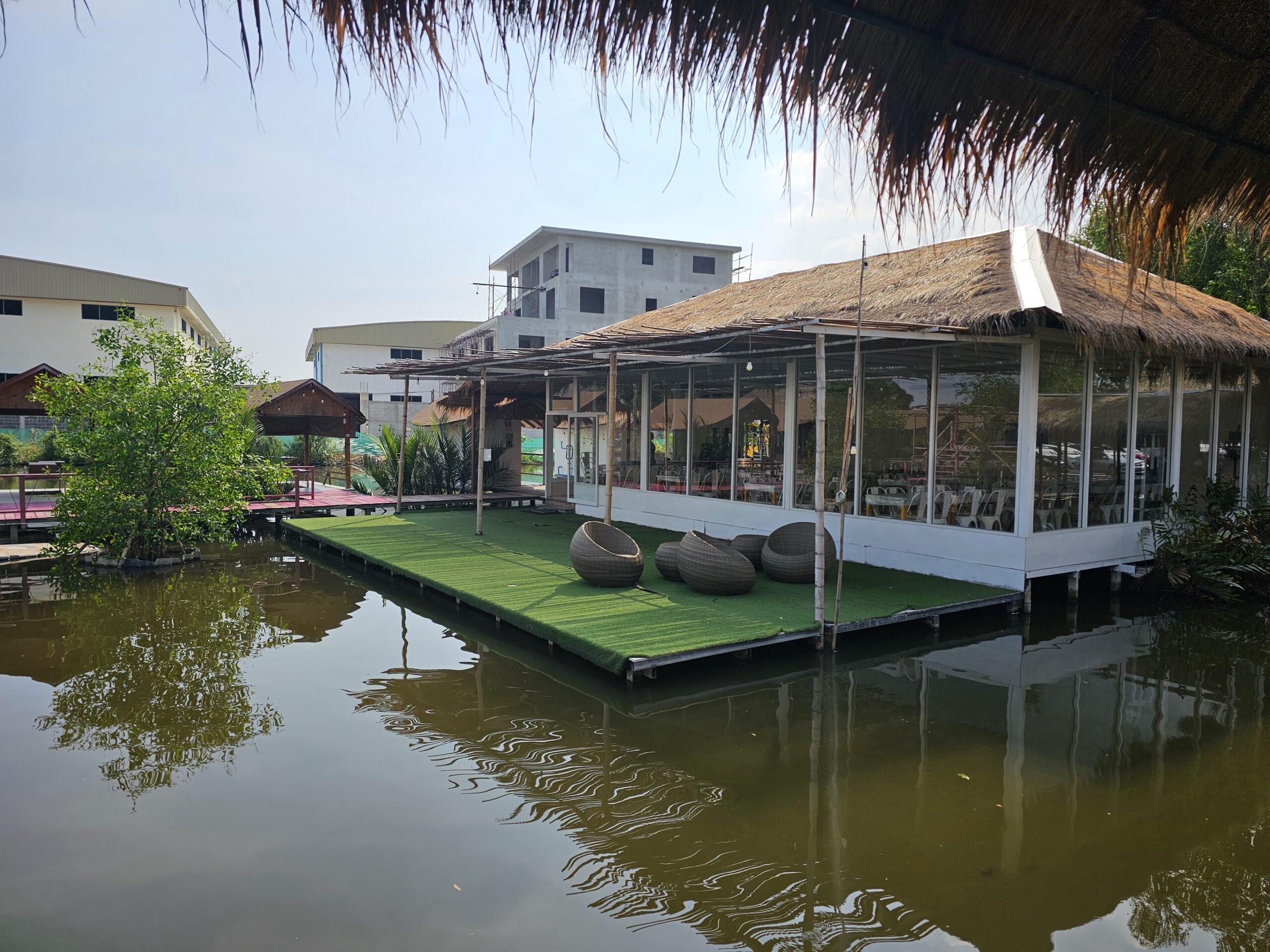 ร้านอาหารลอยน้ำหลังคามุงจากกระจกใสมีเก้าอี้หวายบนสนามหญ้าเทียมริมบ่อน้ำ