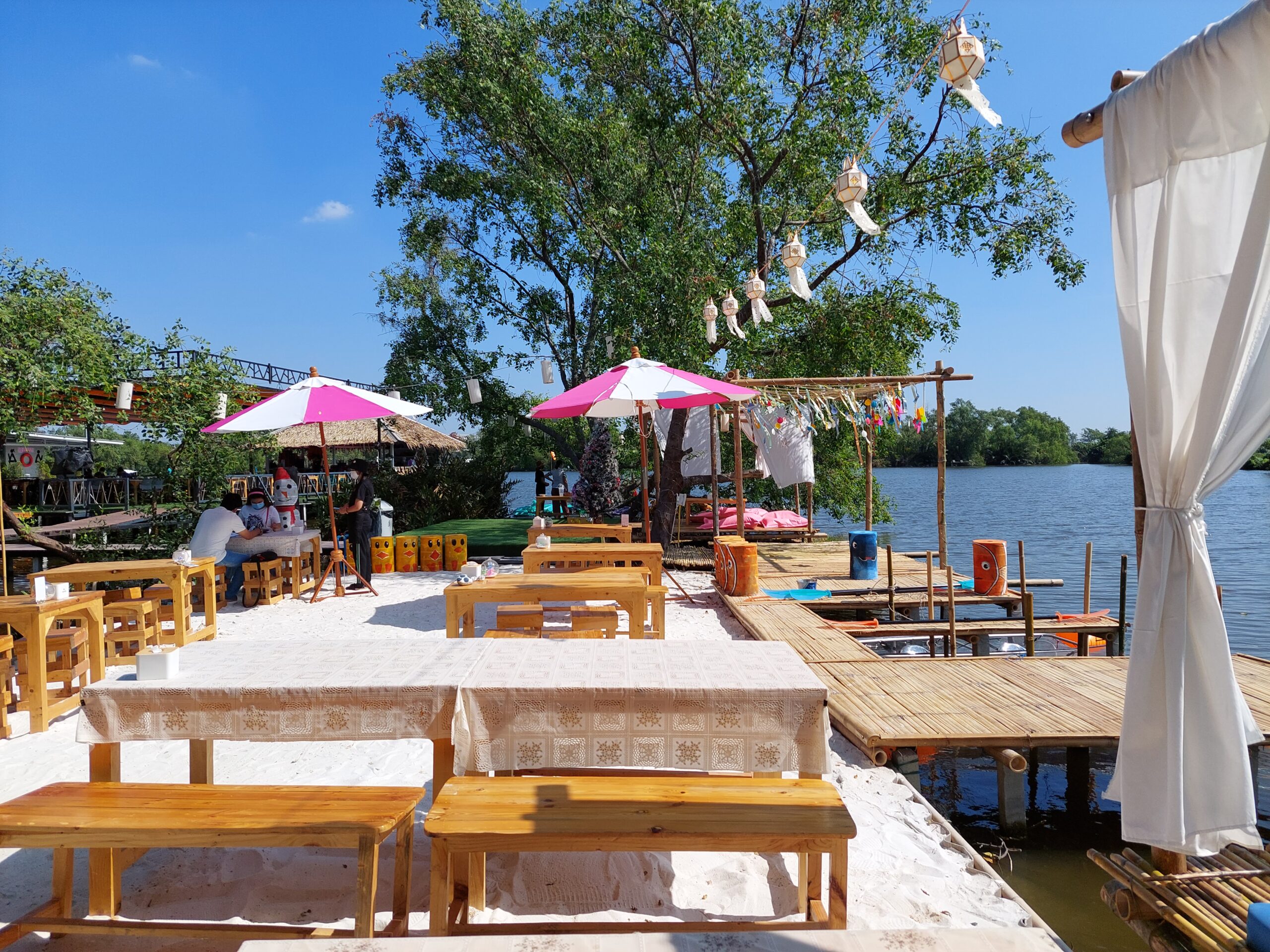 ร้านอาหารริมน้ำบรรยากาศชายหาดทราย มีโต๊ะไม้ ร่มสีชมพู-ขาว แพไม้ไผ่ และโคมไฟตกแต่งริมแม่น้ำ
