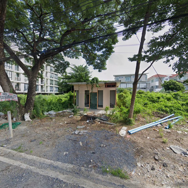 อาคารร้างที่ถูกปกคลุมด้วยพืชพรรณบนที่ดินว่างเปล่าริมถนน มีท่อน้ำสีฟ้าและเศษวัสดุก่อสร้างกระจายอยู่