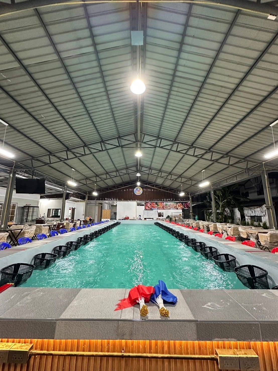 บ่อตกกุ้งในร่มขนาดใหญ่ภายใต้หลังคาเมทัลชีท มีตะกร้าดำเรียงรายรอบบ่อน้ำสีเขียว พร้อมโต๊ะเก้าอี้สำหรับนั่งรับประทานอาหาร