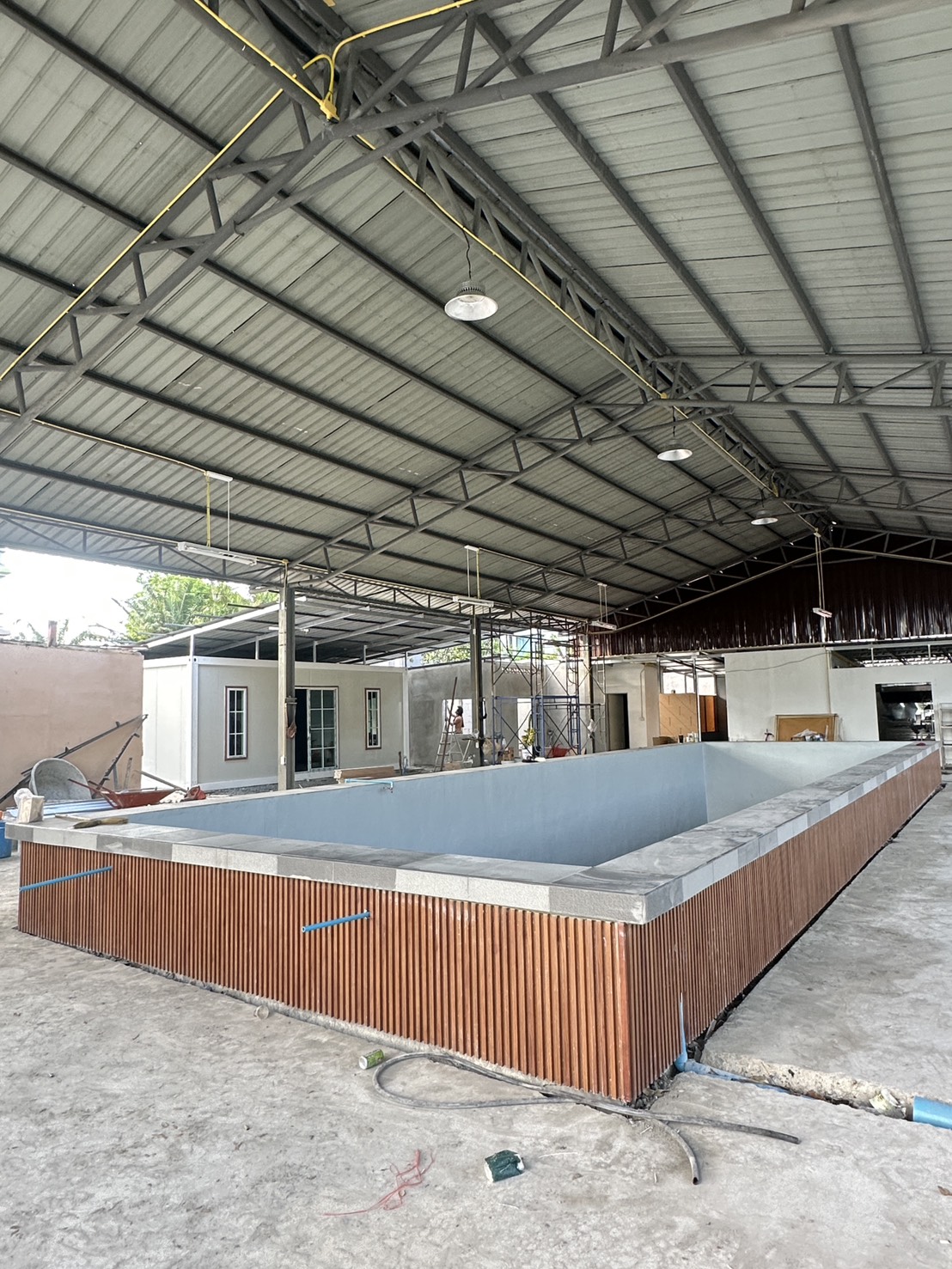 สระว่ายน้ำยกพื้นกำลังก่อสร้างภายใต้หลังคาโครงเหล็ก พร้อมตู้คอนเทนเนอร์สำนักงานและนั่งร้านด้านหลัง