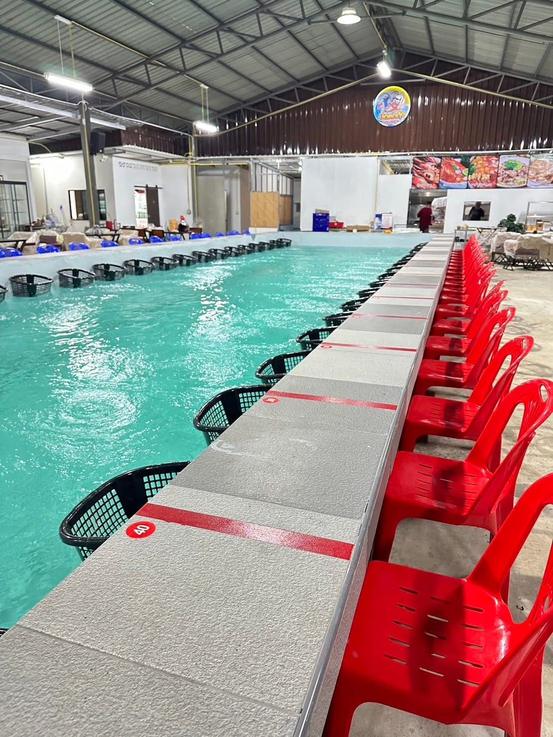 บ่อตกกุ้งในร่มขนาดใหญ่พร้อมเก้าอี้พลาสติกสีแดงเรียงรายและตะกร้าดำริมบ่อน้ำสีเขียว