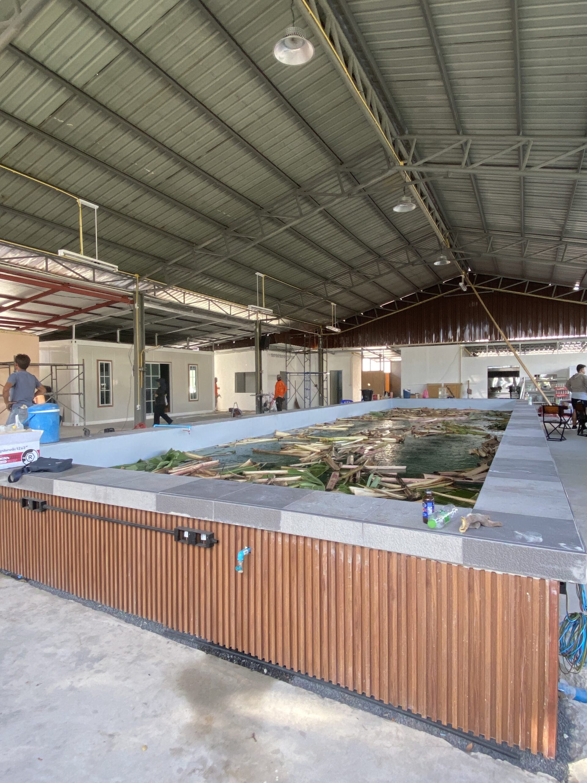 บริเวณก่อสร้างร้านอาหารภายในโกดังหลังคาเหล็ก มีบ่อน้ำตกแต่งด้วยไม้และพื้นกระเบื้อง กำลังอยู่ระหว่างการตกแต่งภายใน