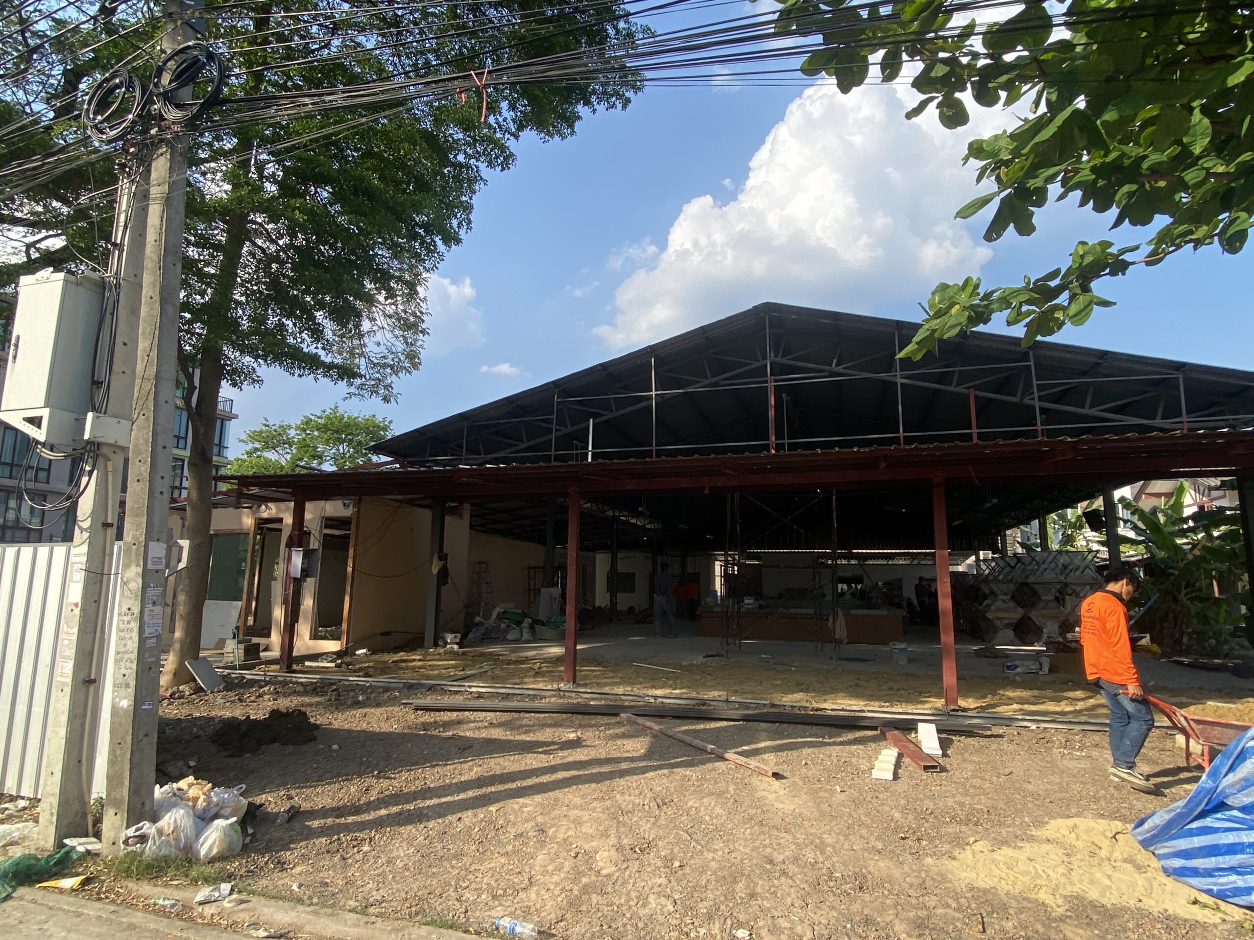 ไซต์ก่อสร้างร้านอาหารที่กำลังสร้างโครงหลังคาเหล็กและเสาเหล็กสีแดง มีคนงานกำลังทำงานอยู่