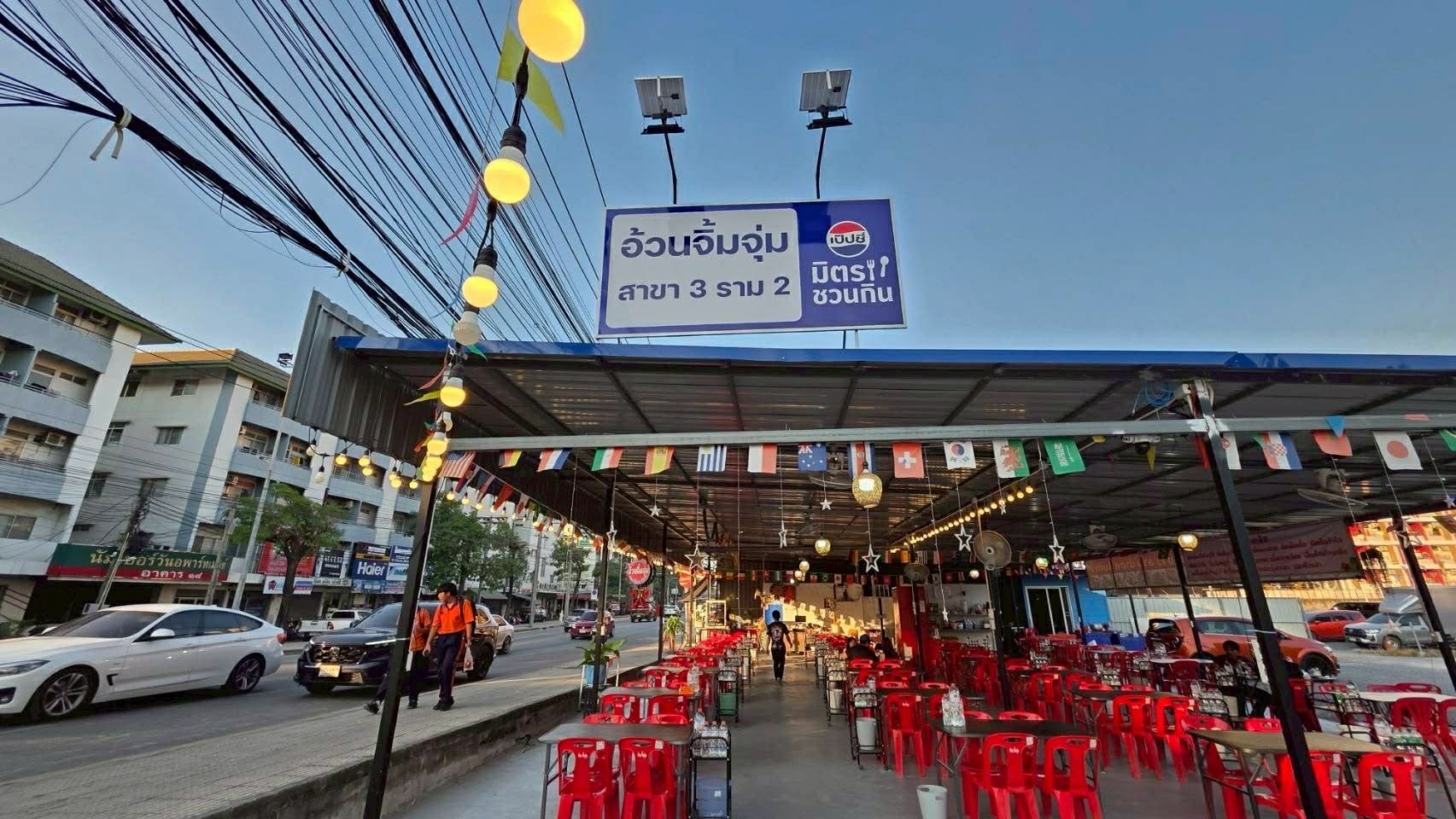 ร้านอ้วนจิ้มจุ่ม สาขา 3 ราม 2 พร้อมโต๊ะเก้าอี้แดงเรียงรายใต้หลังคาเหล็กประดับธงนานาชาติและไฟสายสวยงาม