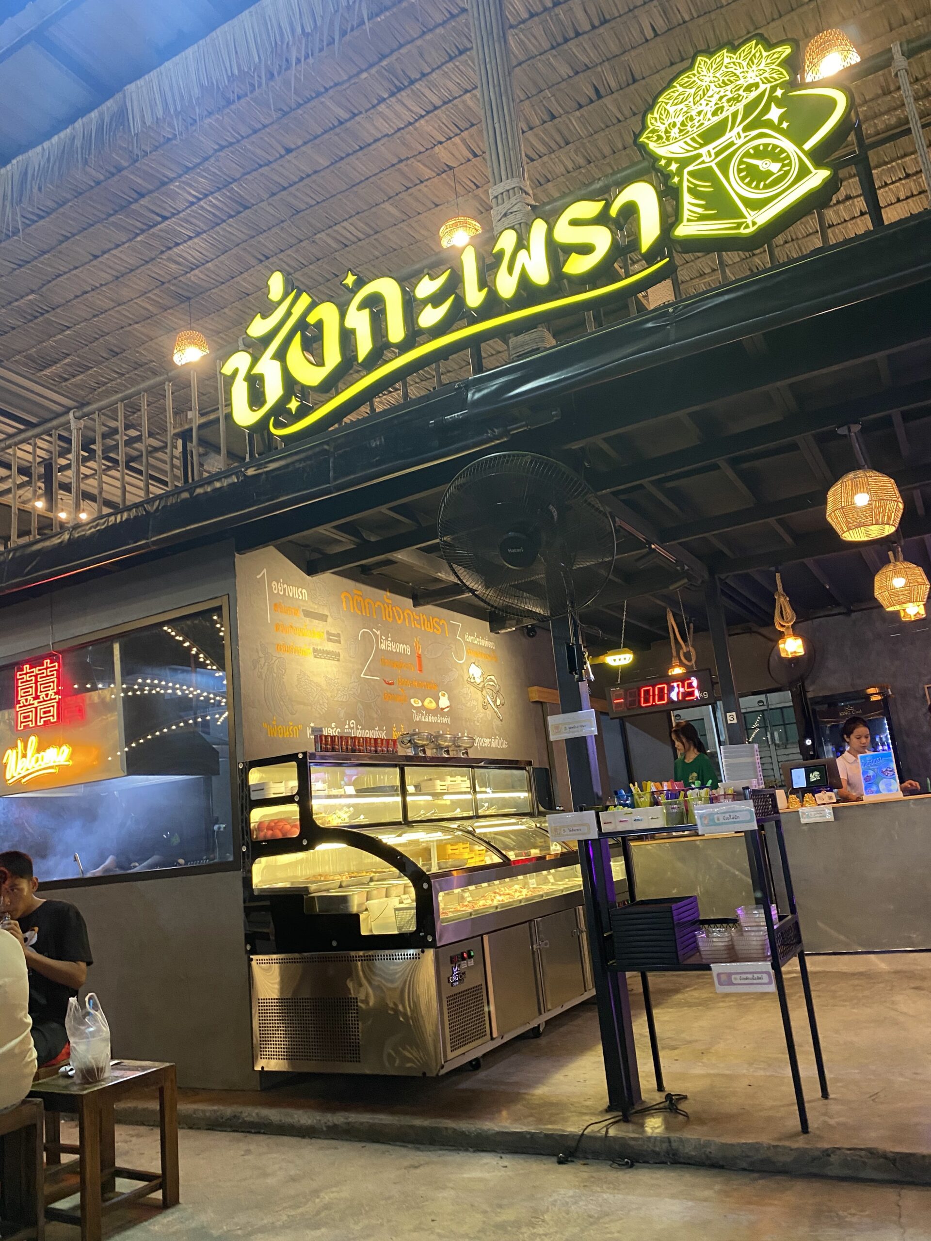 ร้านชั่งกะเพรา บรรยากาศหน้าร้านยามค่ำคืน มีตู้โชว์อาหารและป้ายไฟสีเขียวเหลือง