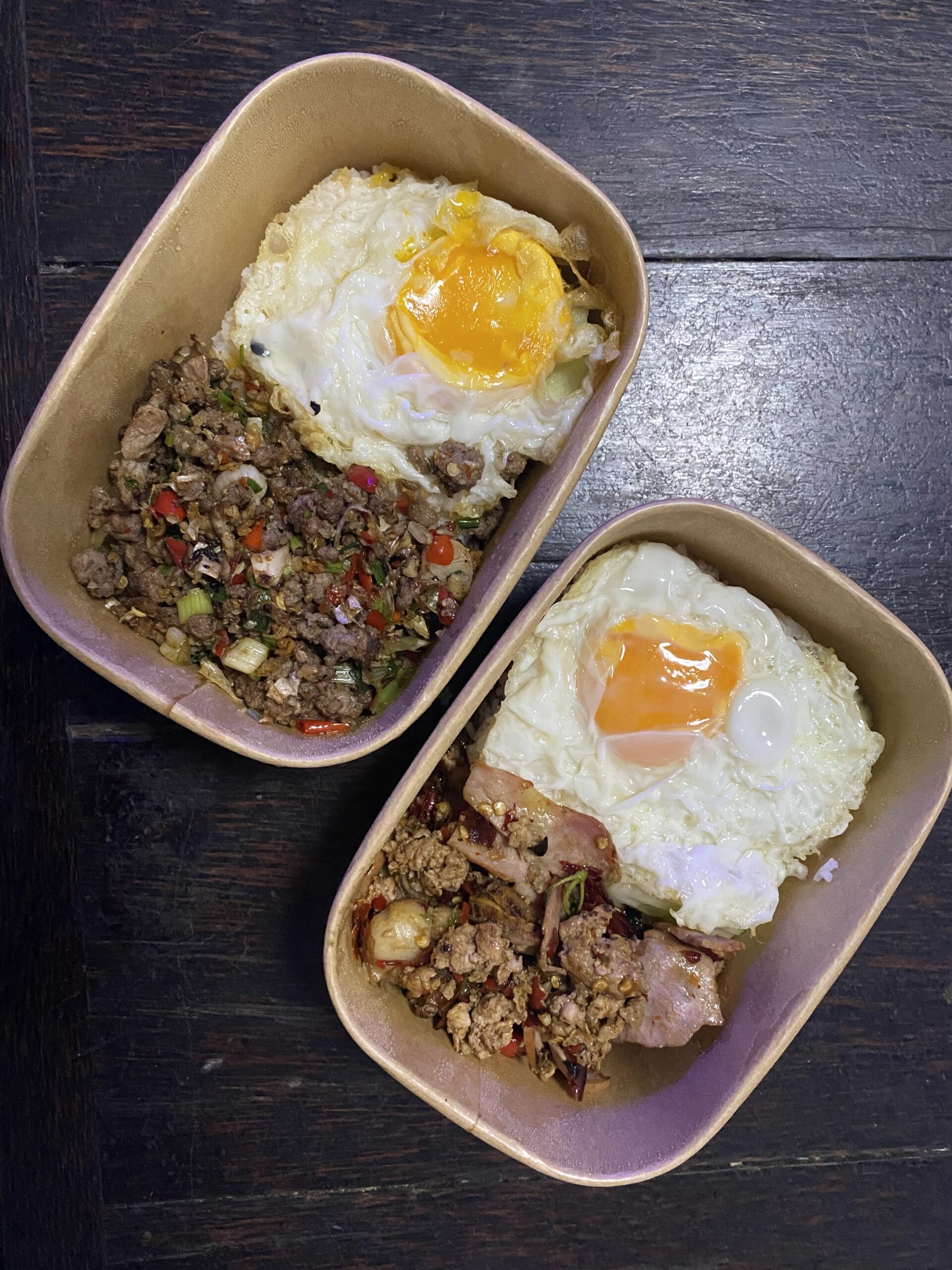 ข้าวกะเพราหมูสับและข้าวกะเพราหมูกรอบไข่ดาวในกล่องกระดาษบนโต๊ะไม้เข้ม