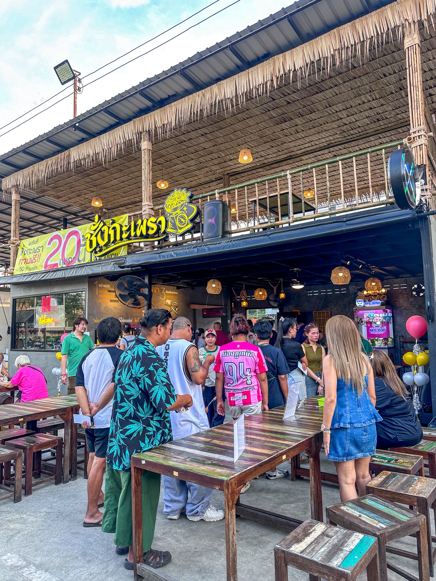 ร้านอาหารไทยชื่อ ชั่งกะเพรา บรรยากาศคึกคัก มีลูกค้ายืนรอเข้าร้านจำนวนมาก โต๊ะไม้เก่าวินเทจ หลังคามุงจาก ป้ายโปรโมชั่น 20 บาท