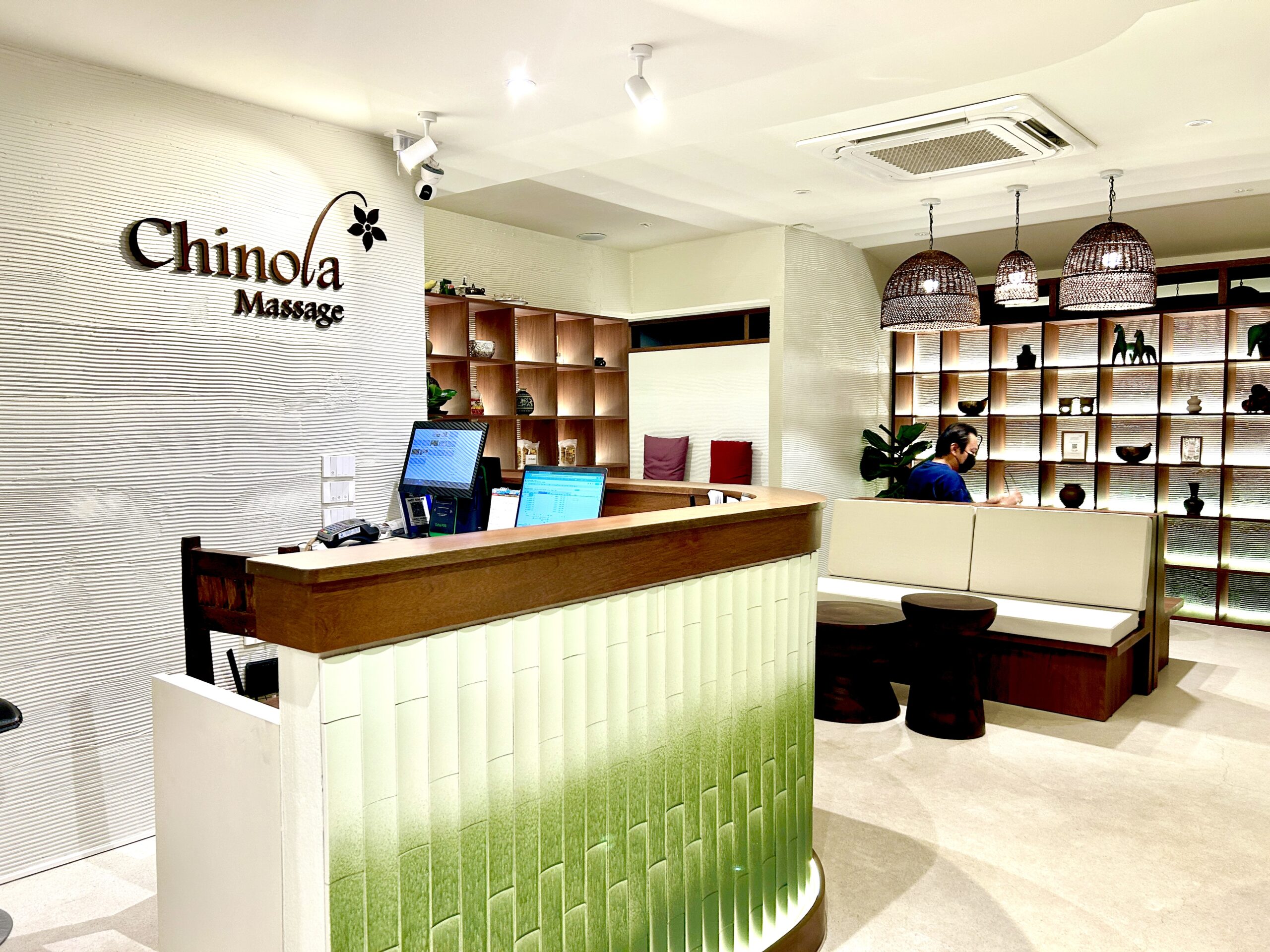 ล็อบบี้ต้อนรับของร้าน Chinola Massage ตกแต่งสวยงามด้วยเคาน์เตอร์สีขาวเขียว ชั้นวางของไม้ และโคมไฟหวาย