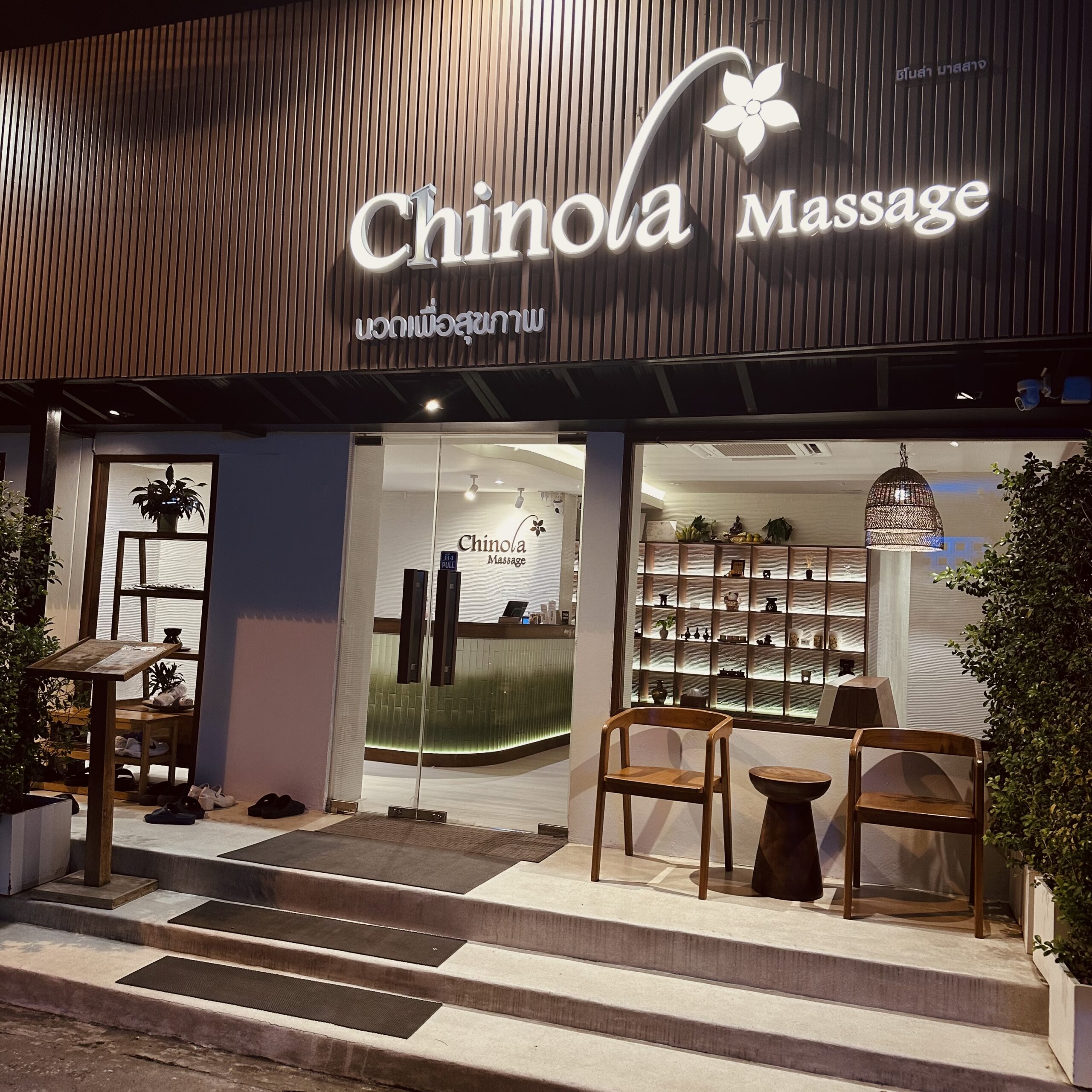 หน้าร้านชิโนล่า มาสสาจ ในเวลากลางคืน มีป้ายไฟสีขาวสว่าง เคาน์เตอร์ต้อนรับ ชั้นวางของตกแต่ง และเก้าอี้ไม้หน้าร้าน