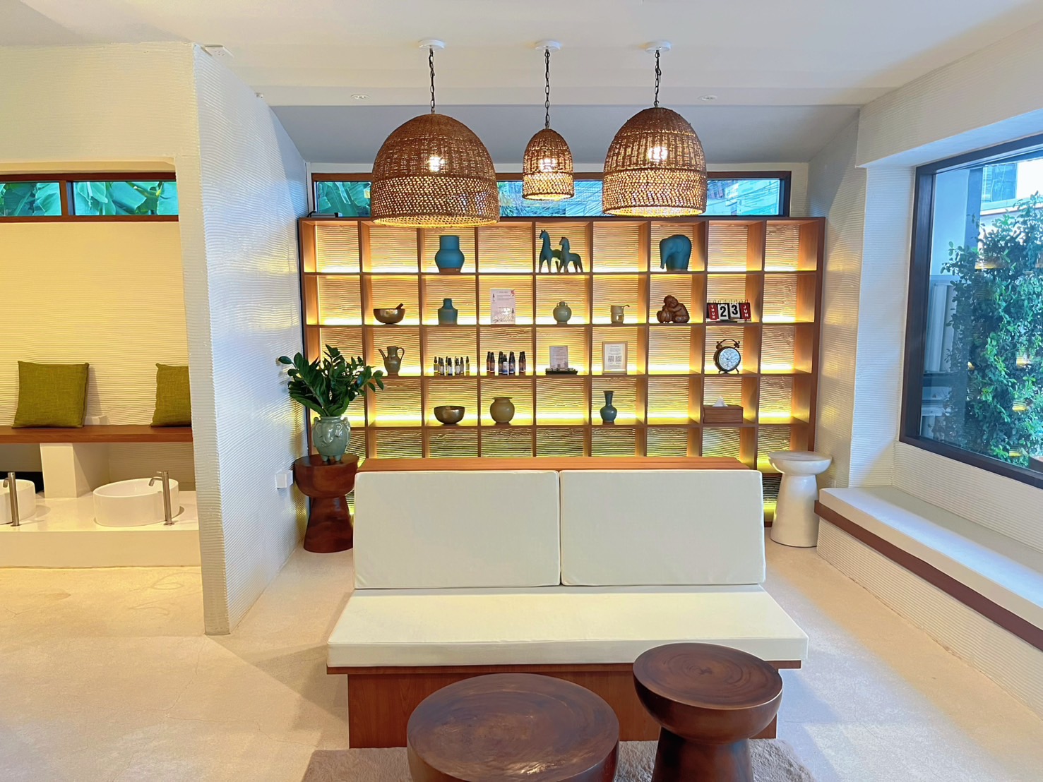 ห้องรับแขกสไตล์โมเดิร์นทรอปิคอลพร้อมโคมไฟหวายแขวนเพดาน ชั้นวางของตกแต่งมีไฟส่อง โซฟาสีขาว และโต๊ะไม้ทรงกลม