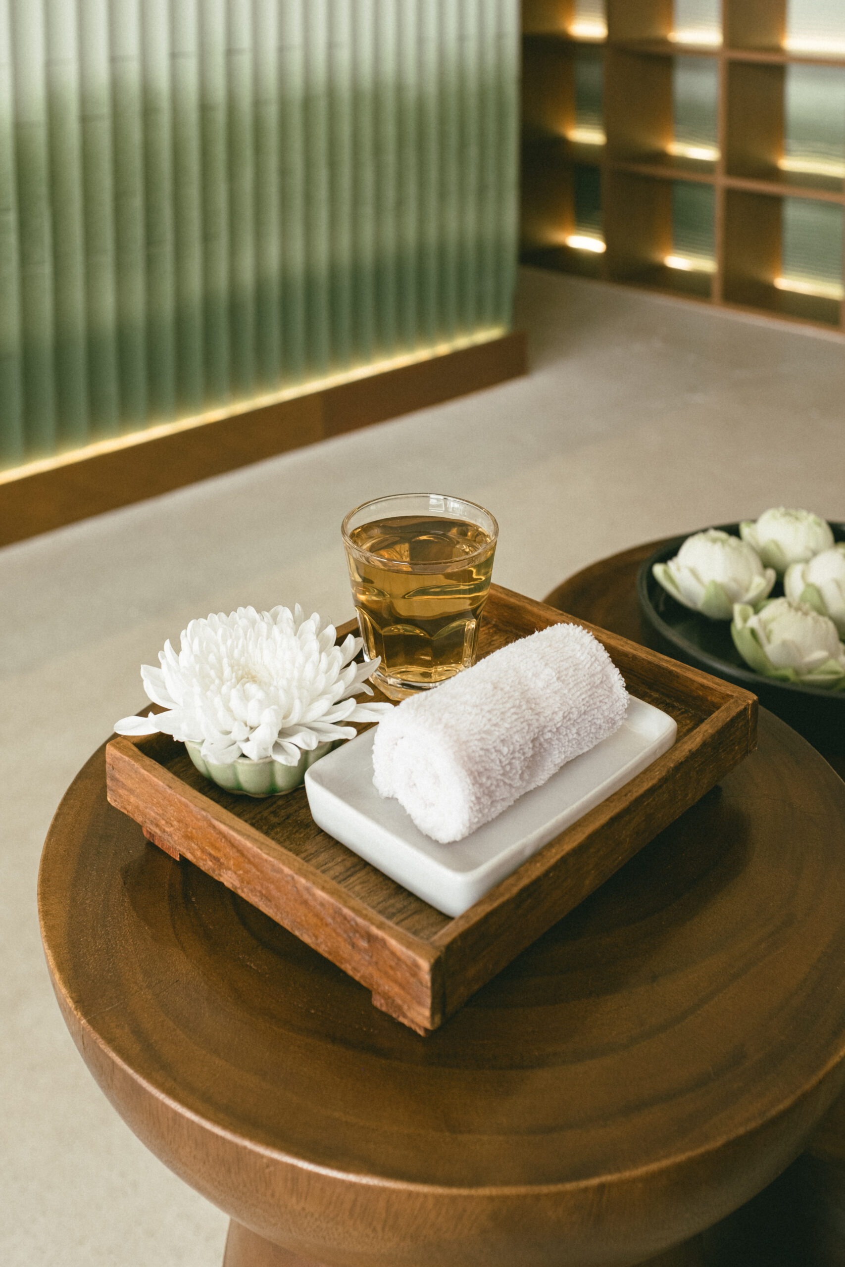 ถาดไม้ต้อมรับแขกพร้อมชาเย็นในแก้วใส ผ้าเย็นม้วนบนจานขาว และดอกเบญจมาศสีขาว บนโต๊ะไม้กลมในบรรยากาศสปาสุดหรู