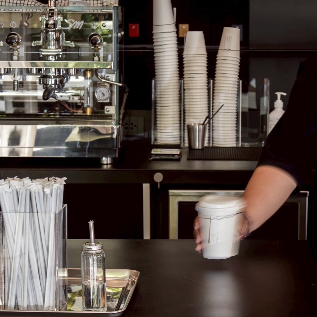 บาริสต้ากำลังเสิร์ฟกาแฟในแก้วกระดาษสีขาวบนเคาน์เตอร์บาร์ พร้อมเครื่องชงกาแฟเอสเพรสโซและแก้วกระดาษซ้อนเรียงอยู่ด้านหลัง
