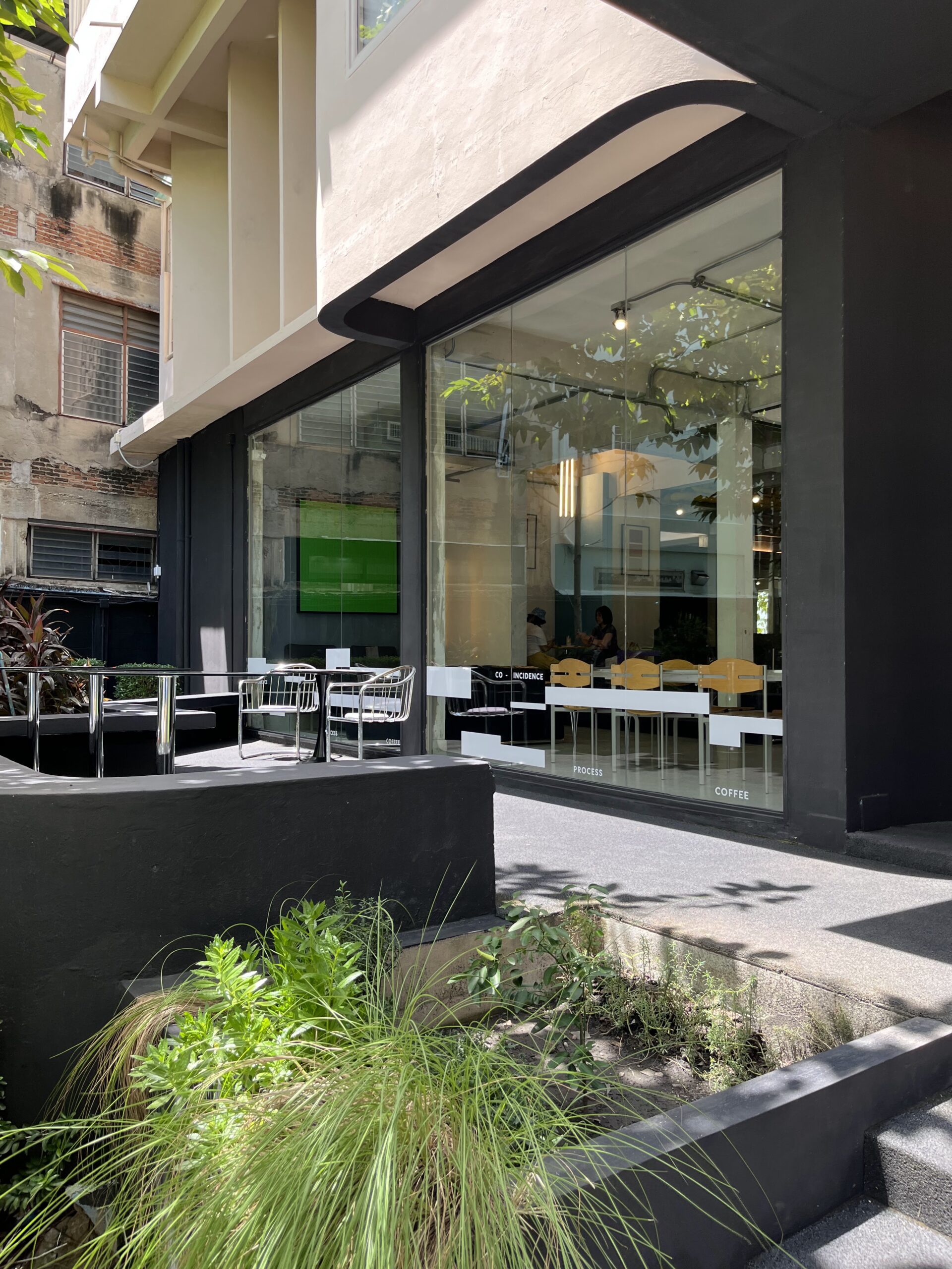 ภายนอกร้านกาแฟ Co-Incidence Process Coffee ที่มีกระจกบานใหญ่ โต๊ะนั่งกลางแจ้ง และสวนหย่อมสีเขียว