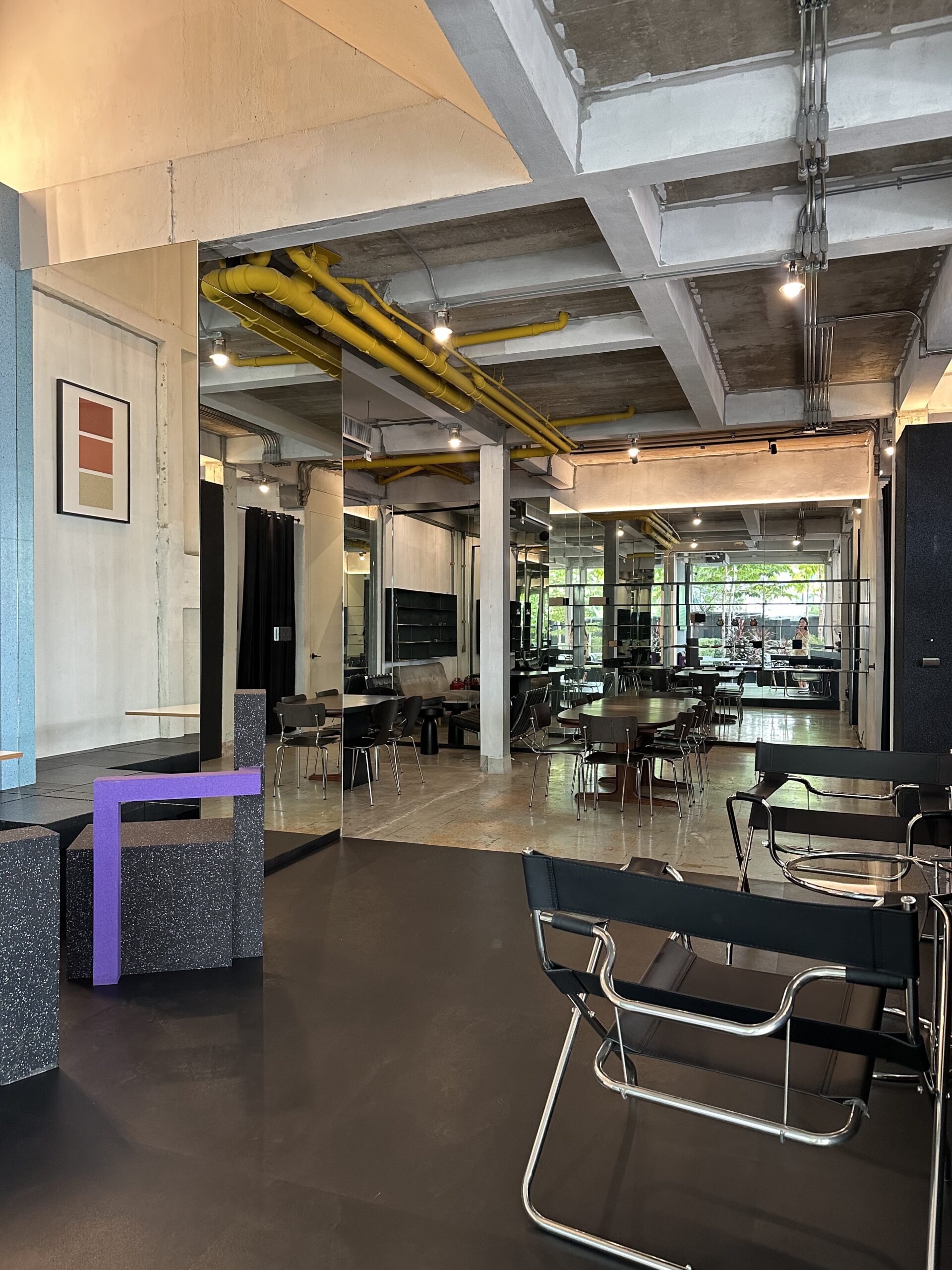 ภายในร้านอาหารสไตล์ลอฟต์อินดัสเทรียล มีเพดานคอนกรีตเปลือย ท่อสีเหลือง เก้าอี้หนังสีดำแบบ Wassily และเฟอร์นิเจอร์สีม่วงกับกระจกบานใหญ่