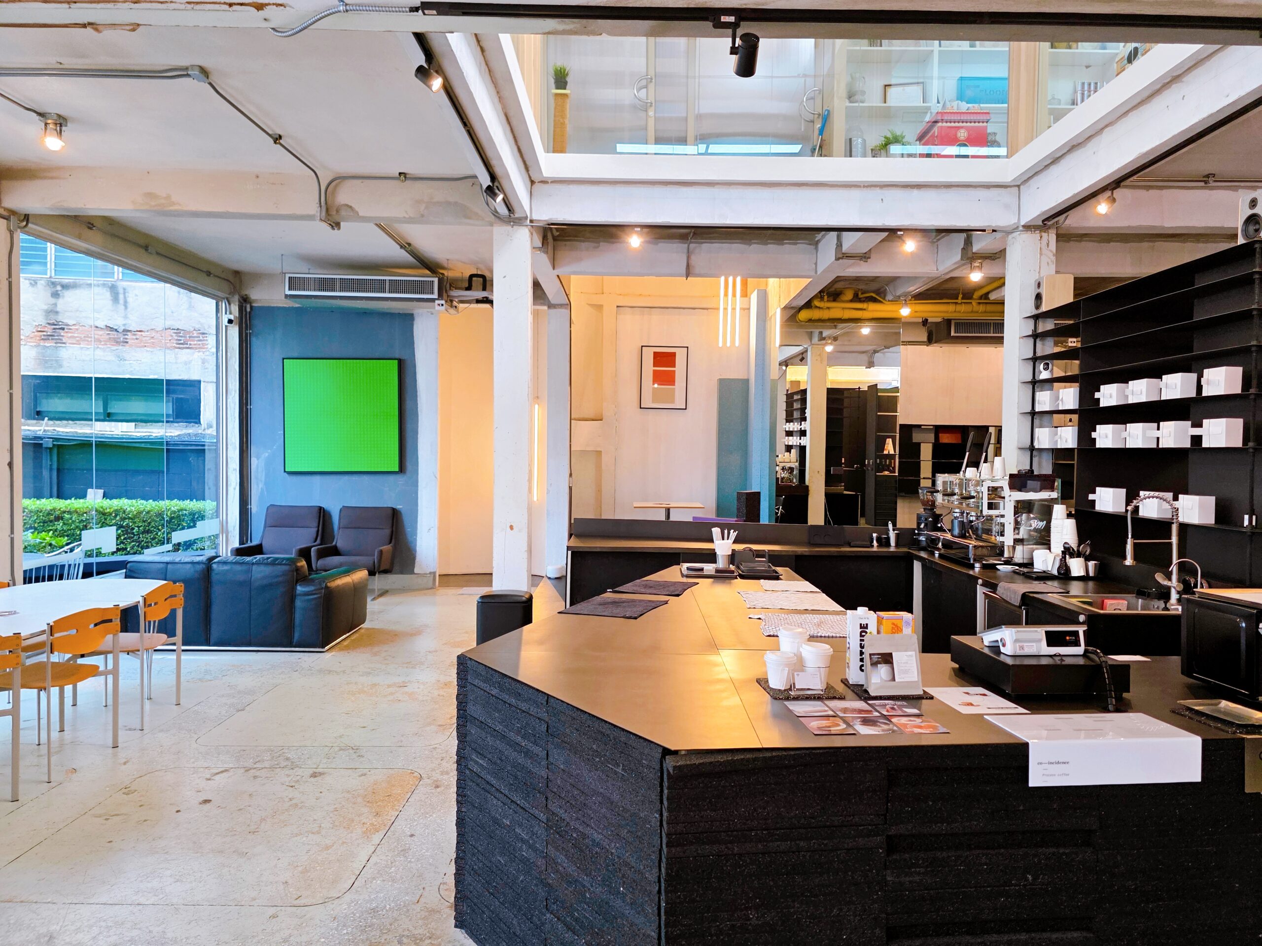 ภายในร้านกาแฟสไตล์อินดัสเทรียลพร้อมเคาน์เตอร์บาร์สีดำ โซฟานั่งเล่นสีน้ำเงินเข้ม และผนังสีฟ้าพร้อมจอสีเขียว