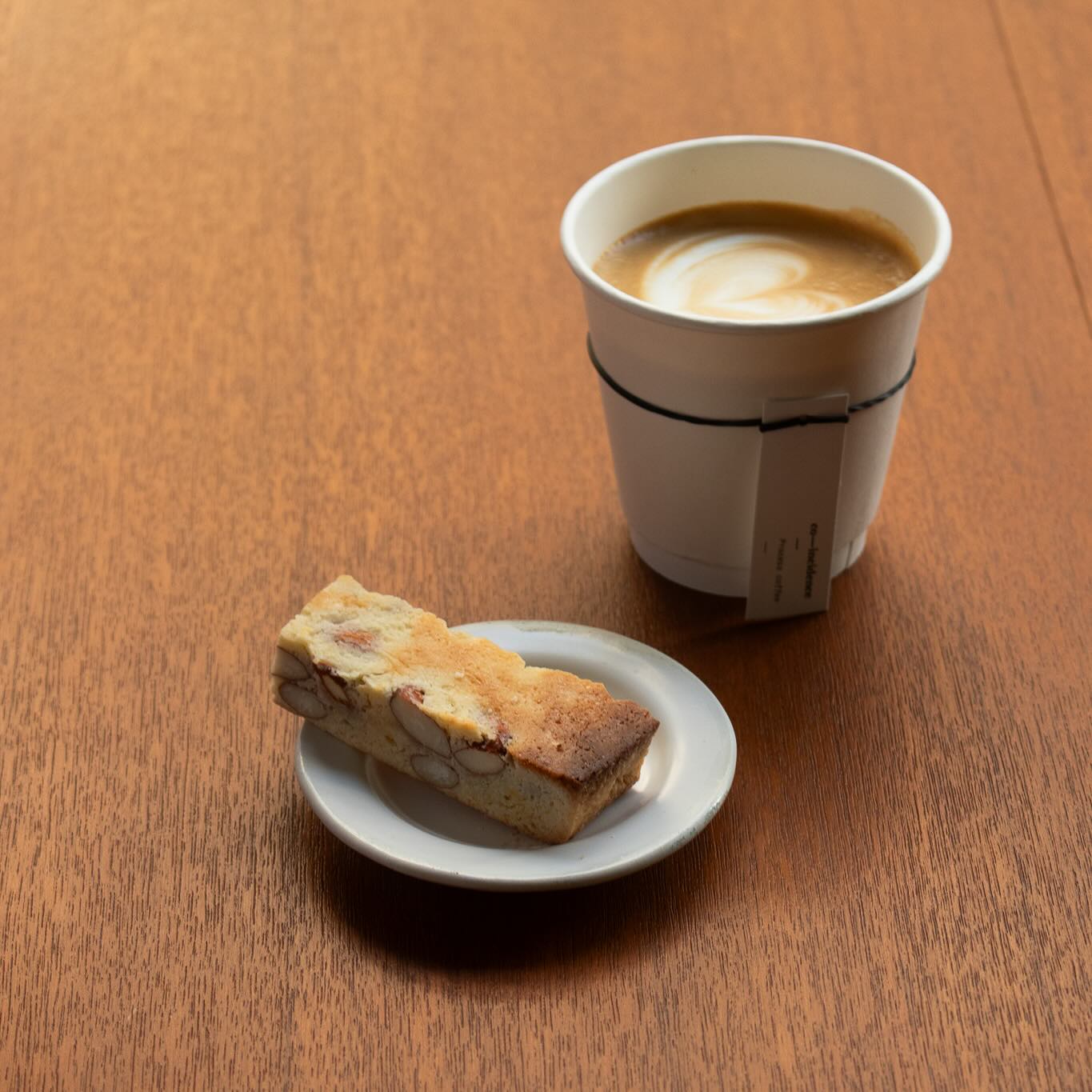 กาแฟลาเต้อาร์ตในแก้วกระดาษพร้อมบิสคอตตี้อัลมอนด์บนจานเล็กวางบนโต๊ะไม้