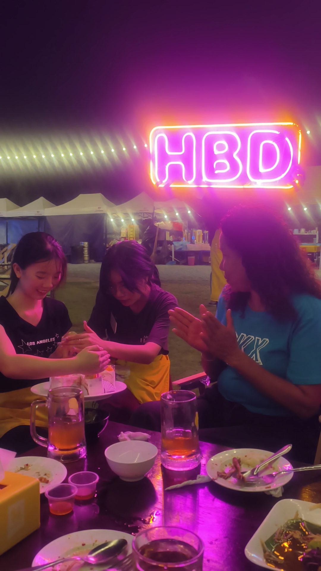 กลุ่มเพื่อนสาวนั่งฉลองวันเกิดที่ร้านอาหารกลางแจ้งมีป้ายไฟนีออน HBD สีชมพูม่วง พร้อมอาหารและเครื่องดื่มบนโต๊ะ