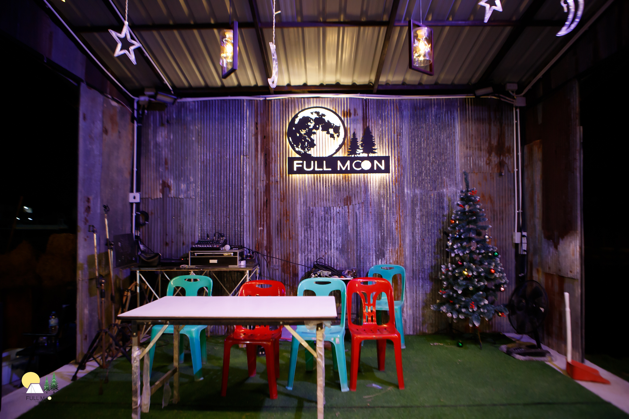พื้นที่นั่งเล่นร้าน Full Moon ตกแต่งด้วยผนังสังกะสี มีโต๊ะเก้าอี้พลาสติกสีแดงและสีเขียว ต้นคริสต์มาส และอุปกรณ์ดีเจ บนพื้นหญ้าเทียมสีเขียว
