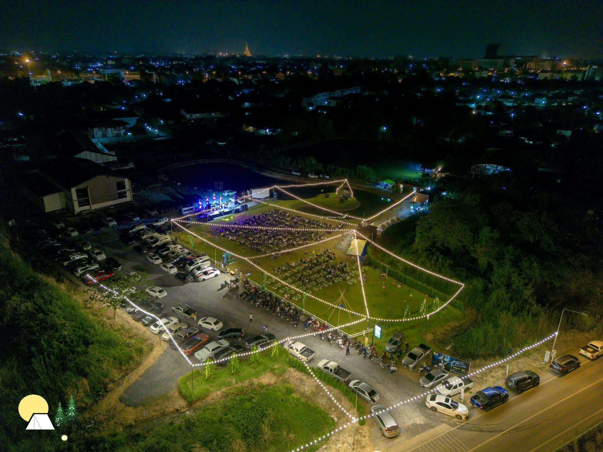 ภาพถ่ายมุมสูงของสถานที่จัดงานกลางแจ้งในเวลากลางคืน มีเวทีคอนเสิร์ต ไฟประดับสายสวยงาม ลานจอดรถ และผู้คนนั่งชมการแสดงบนสนามหญ้า มีวิวเมืองเป็นฉากหลัง