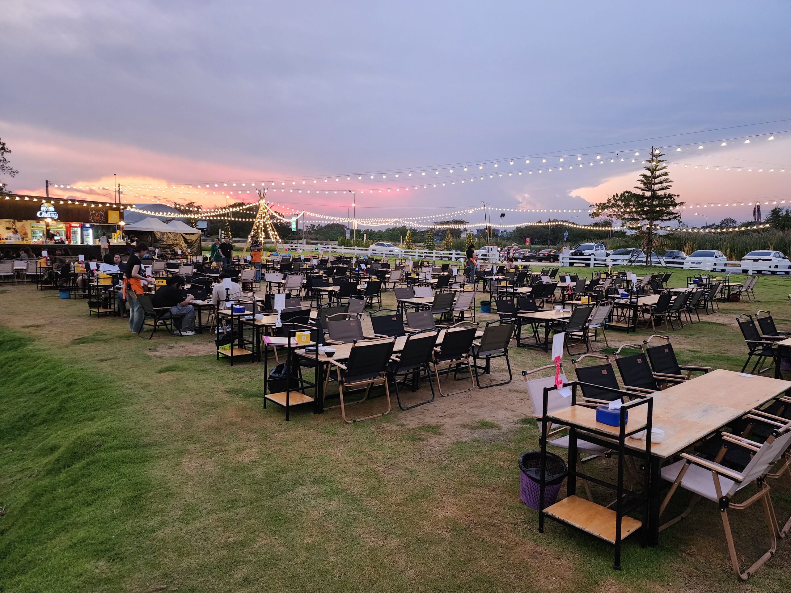 ร้านอาหารกลางแจ้งบนสนามหญ้าพร้อมโต๊ะเก้าอี้แคมป์ปิ้งจำนวนมาก ตกแต่งด้วยไฟสายประดับและกระโจมไฟในยามพระอาทิตย์ตก มีป้ายเบียร์ช้างที่บาร์