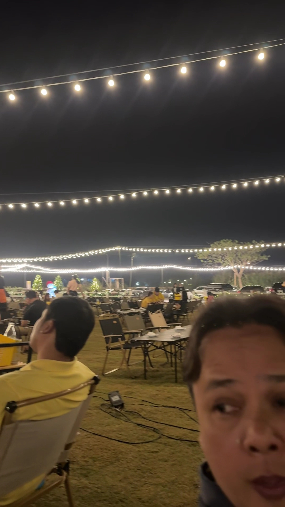 บรรยากาศร้านอาหารกลางแจ้งยามค่ำคืน มีไฟสายประดับสวยงาม โต๊ะเก้าอี้จัดเรียงบนสนามหญ้า มีผู้คนนั่งรับประทานอาหาร