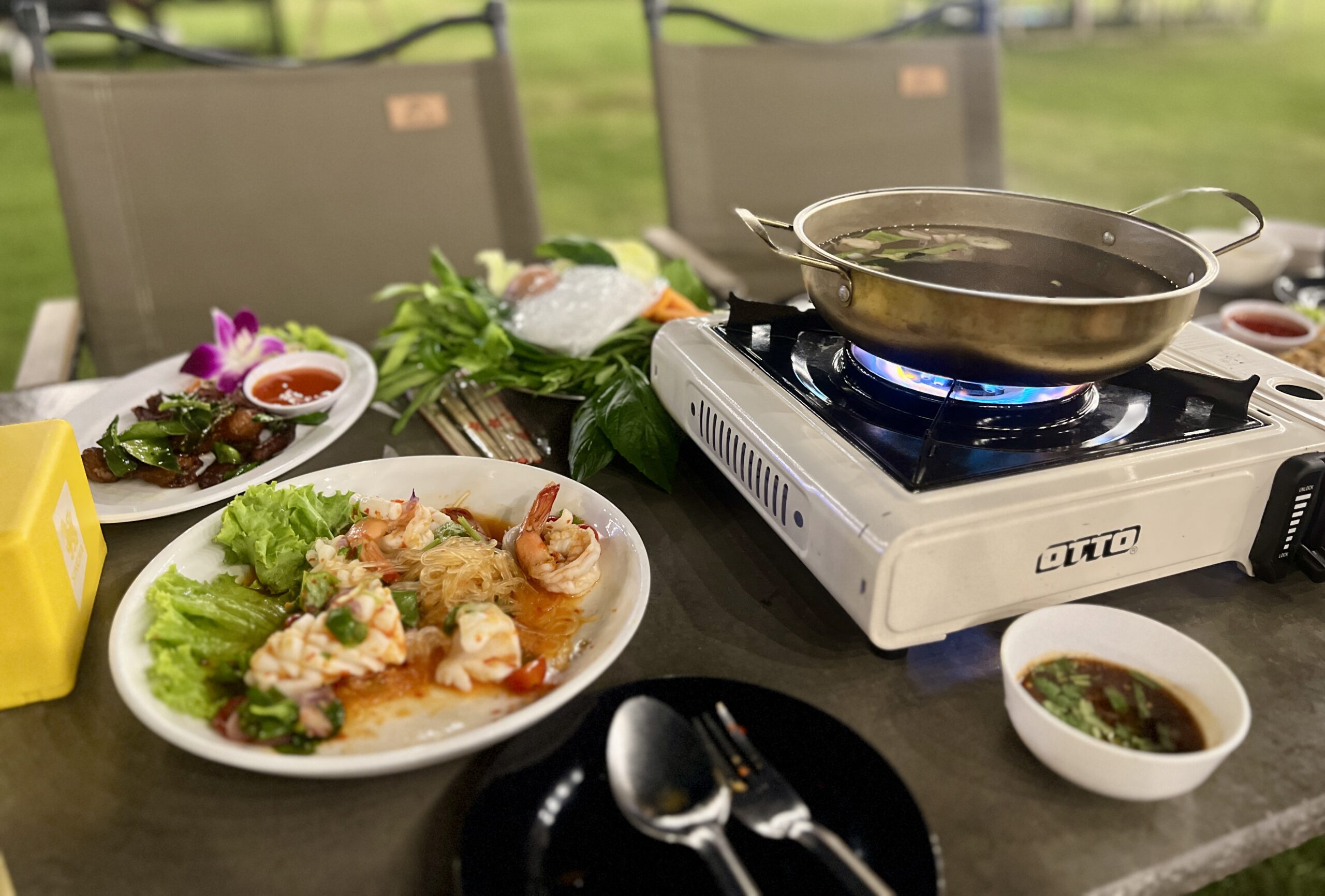 โต๊ะอาหารกลางแจ้งพร้อมเตาแก๊สพกหมู่ยี่ห้อ OTTO กระทะหม้อไฟ จานยำกุ้งสด จานเนื้อทอดกระเทียมพริกไทย ผักสด และน้ำจิ้ม บนพื้นหญ้าสีเขียว