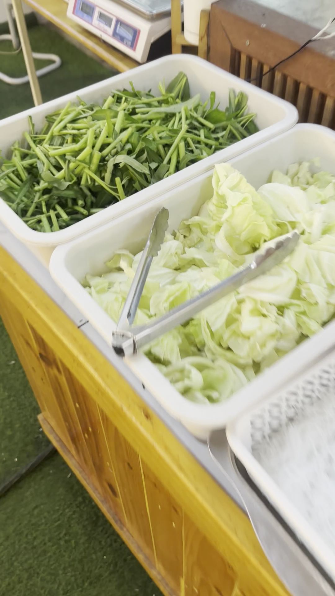 บุฟเฟ่ต์ผักสดมีผักบุ้งและกะหล่ำปลีหั่นในถาดสีขาวพร้อมที่คีบสำหรับใส่หม้อไฟ