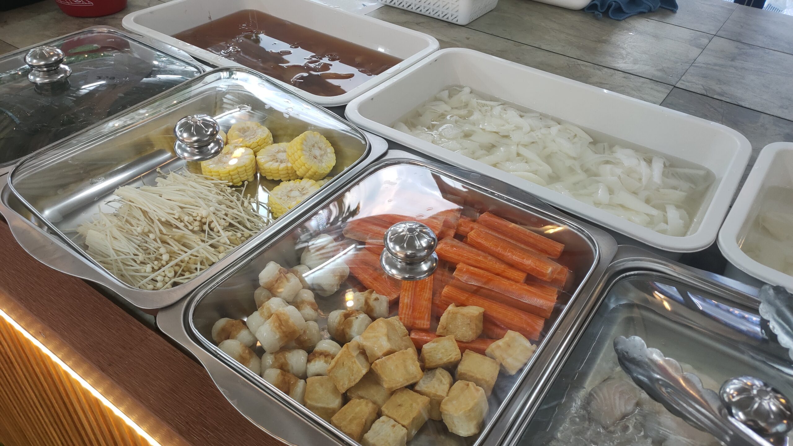 บุฟเฟ่ต์ชาบูพร้อมวัตถุดิบหลากหลาย เห็ดเข็มทอง ข้าวโพด ปูอัด เต้าหู้ทอด ลูกชิ้น และเส้นก๋วยเตี๋ยวในถาดสแตนเลส