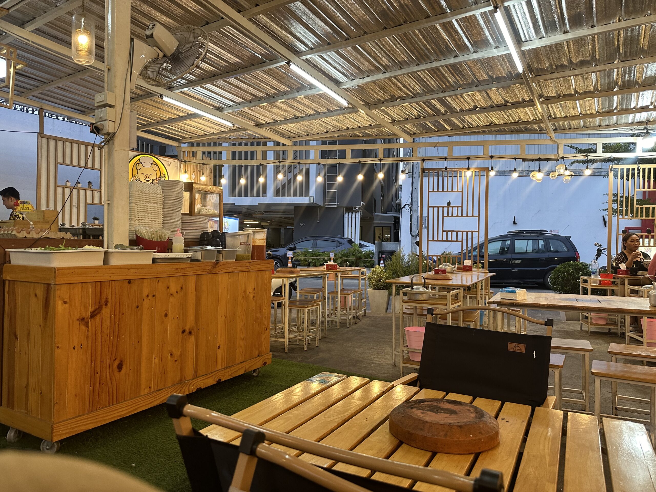 ร้านอาหารเปิดโล่งสไตล์สตรีทฟู้ดมีหลังคาสังกะสี โต๊ะเก้าอี้ไม้ เคาน์เตอร์ไม้สำหรับปรุงอาหาร มีป้ายหมูการ์ตูน สายไฟประดับ และแล็ปท็อปบนโต๊ะ