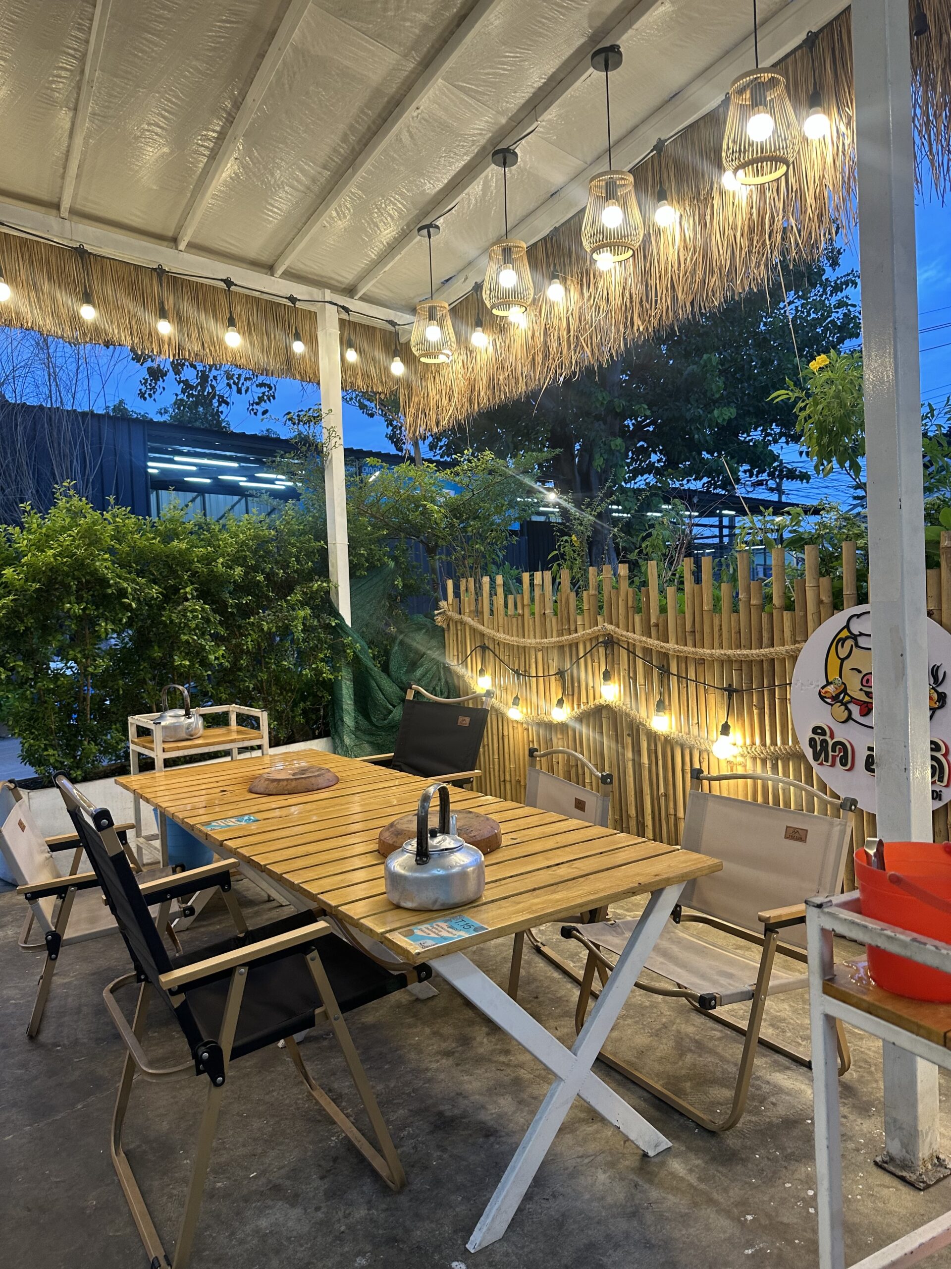 โซนนั่งกลางแจ้งของร้านอาหาร มีโต๊ะไม้พับได้ เก้าอี้แคมป์ปิ้ง รั้วไม้ไผ่ โคมไฟหวาย และไฟประดับยามเย็น