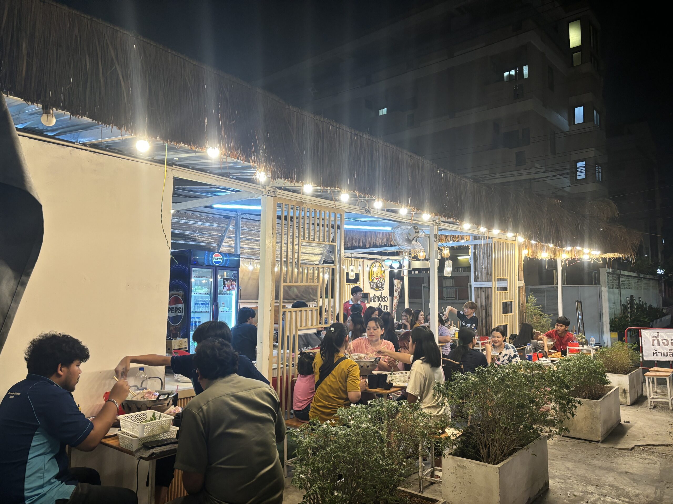 ร้านอาหารกลางแจ้งยามค่ำคืนในประเทศไทย มีลูกค้านั่งทานอาหารเต็มร้าน ตกแต่งด้วยหลังคามุงจากและไฟสายประดับ มีตู้เย็นเป๊ปซี่อยู่ด้านข้าง